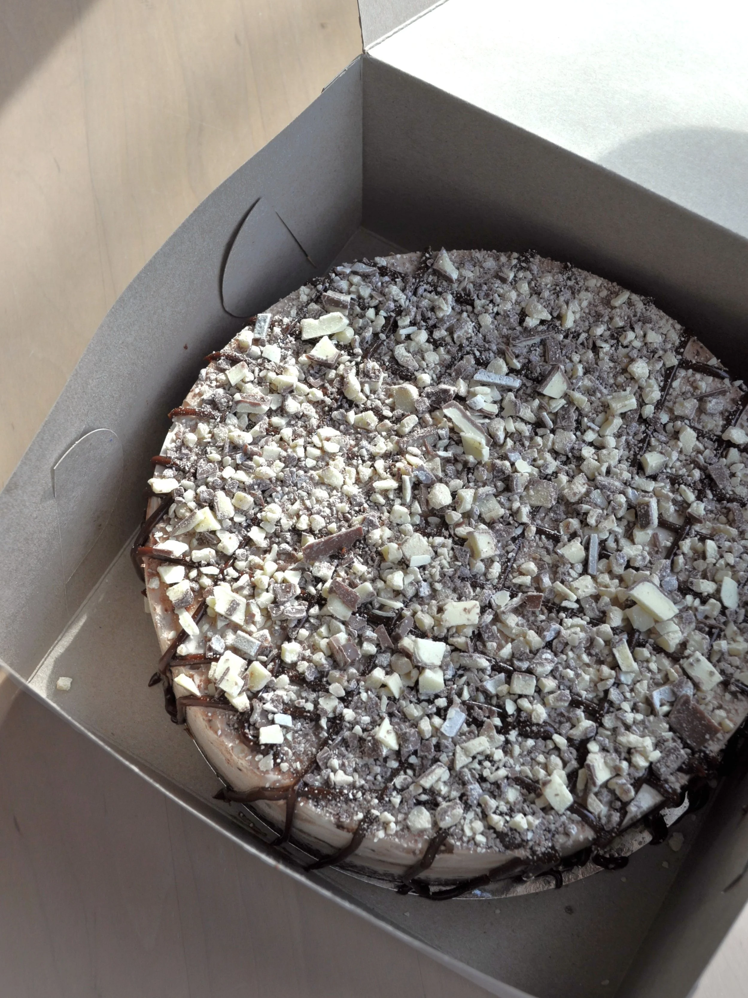 A chocolate ice cream cake with white chocolate and chocolate shavings inside a cake box.