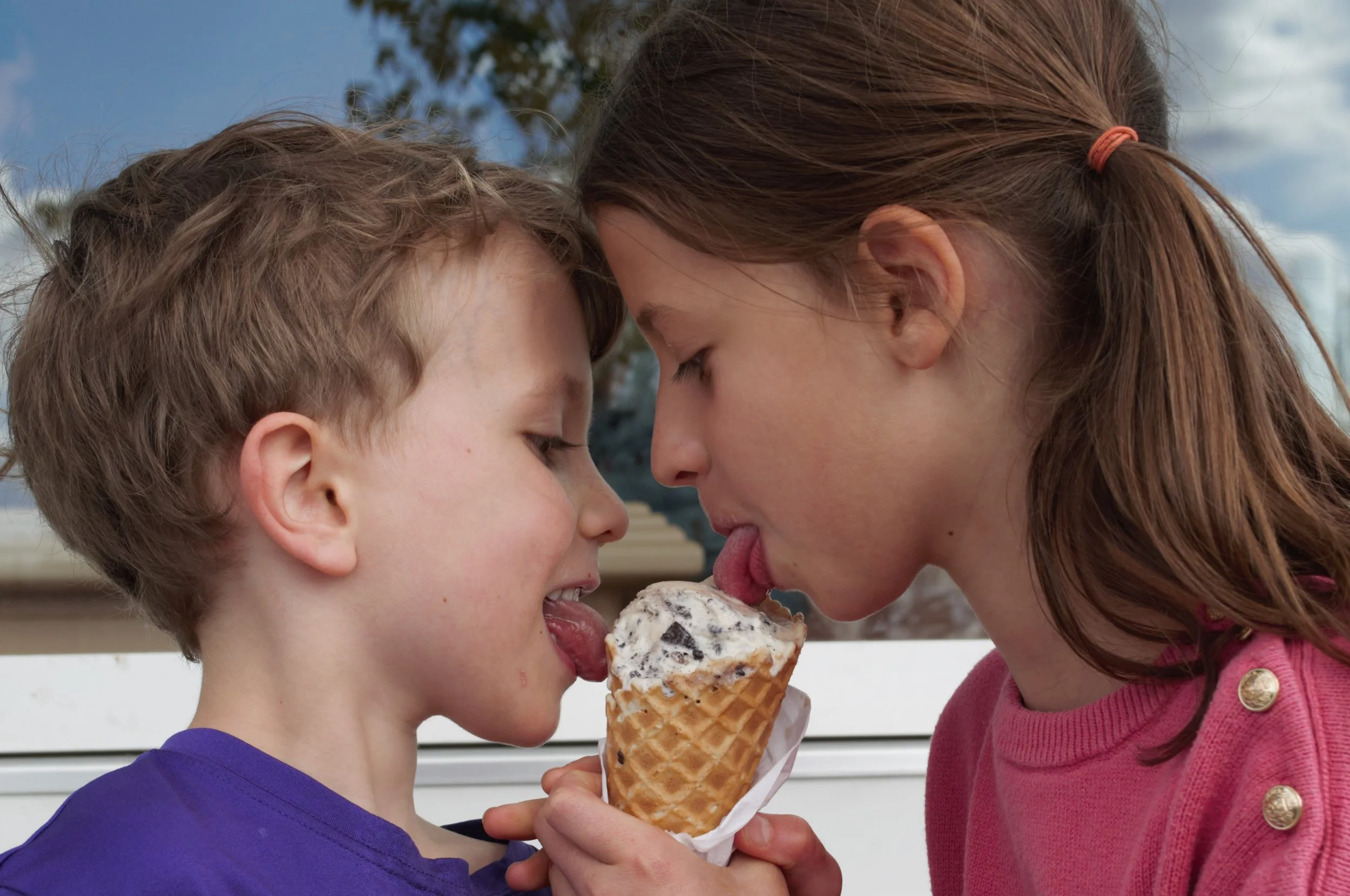 A young boy and girl are sharing a large ice cream cone together, with their foreheads touching and eyes closed, outdoors on a summer day.