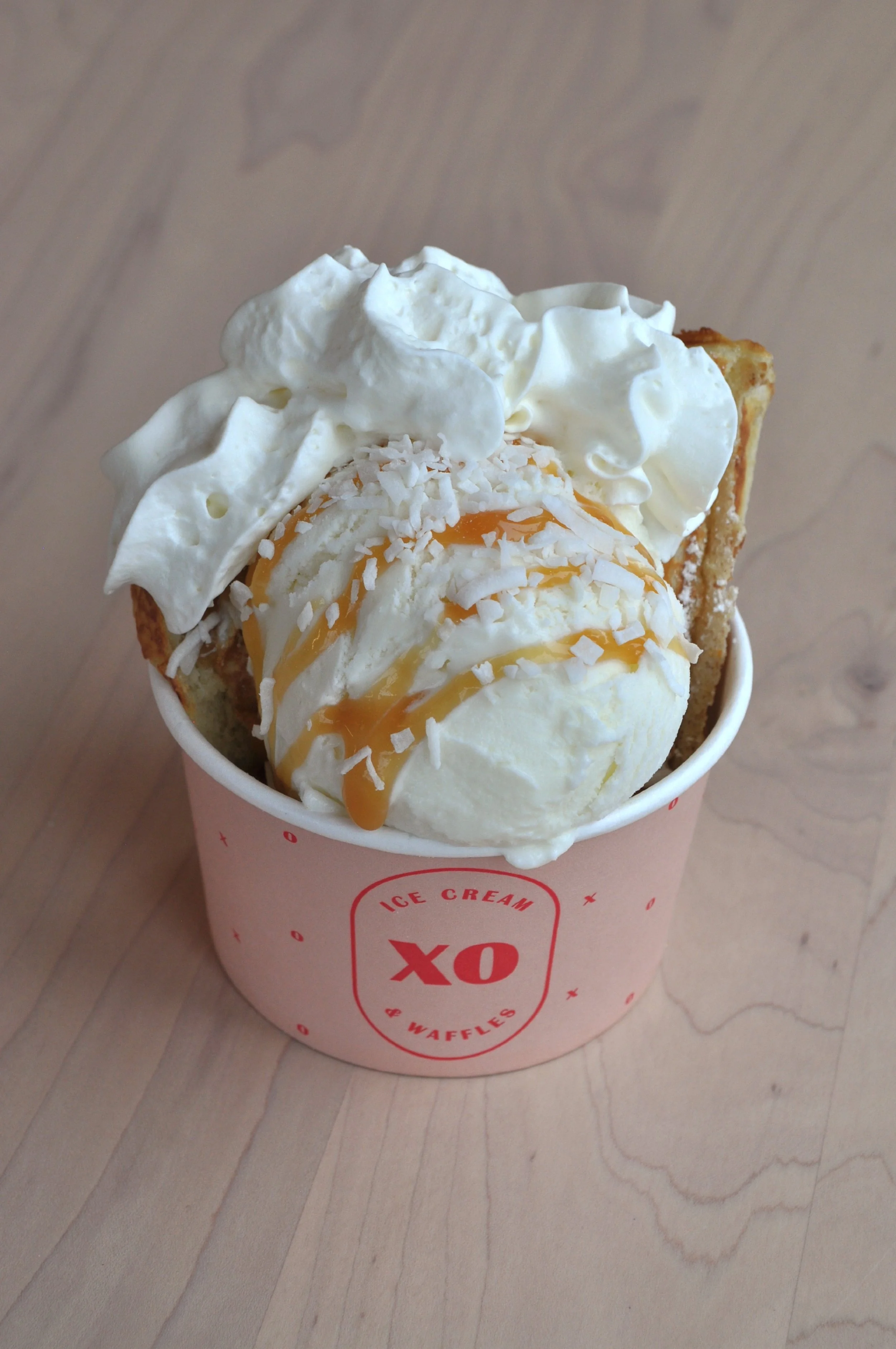 A waffle sundae in a pink cup containing a scoop of ice cream topped with whipped cream, caramel drizzle, shredded coconut and fresh waffle strips.