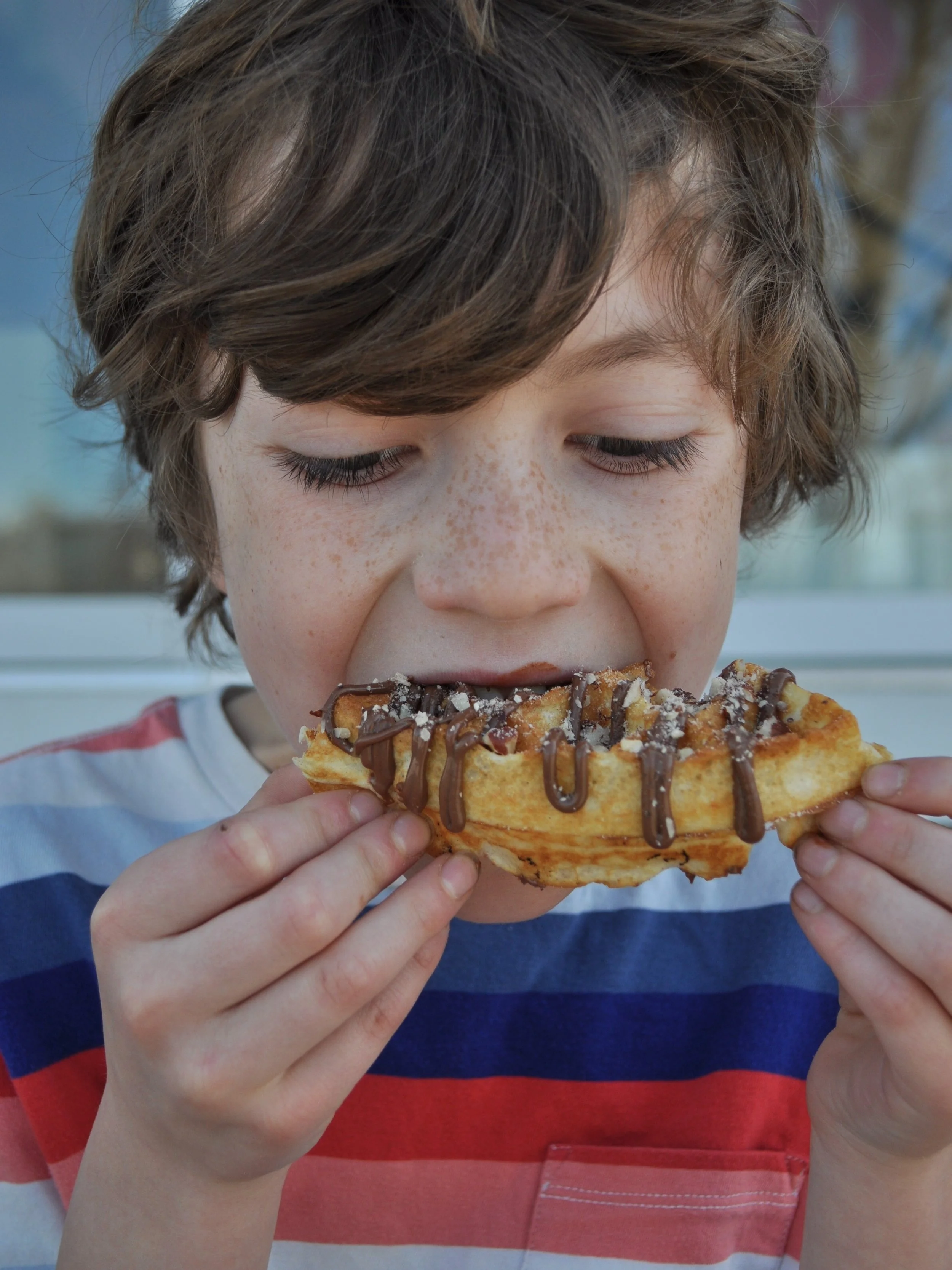A young boy with brown hair and freckles eating a waffle topped with Nutella and chopped hazelnuts. His fingers are getting messy with the Nutella.