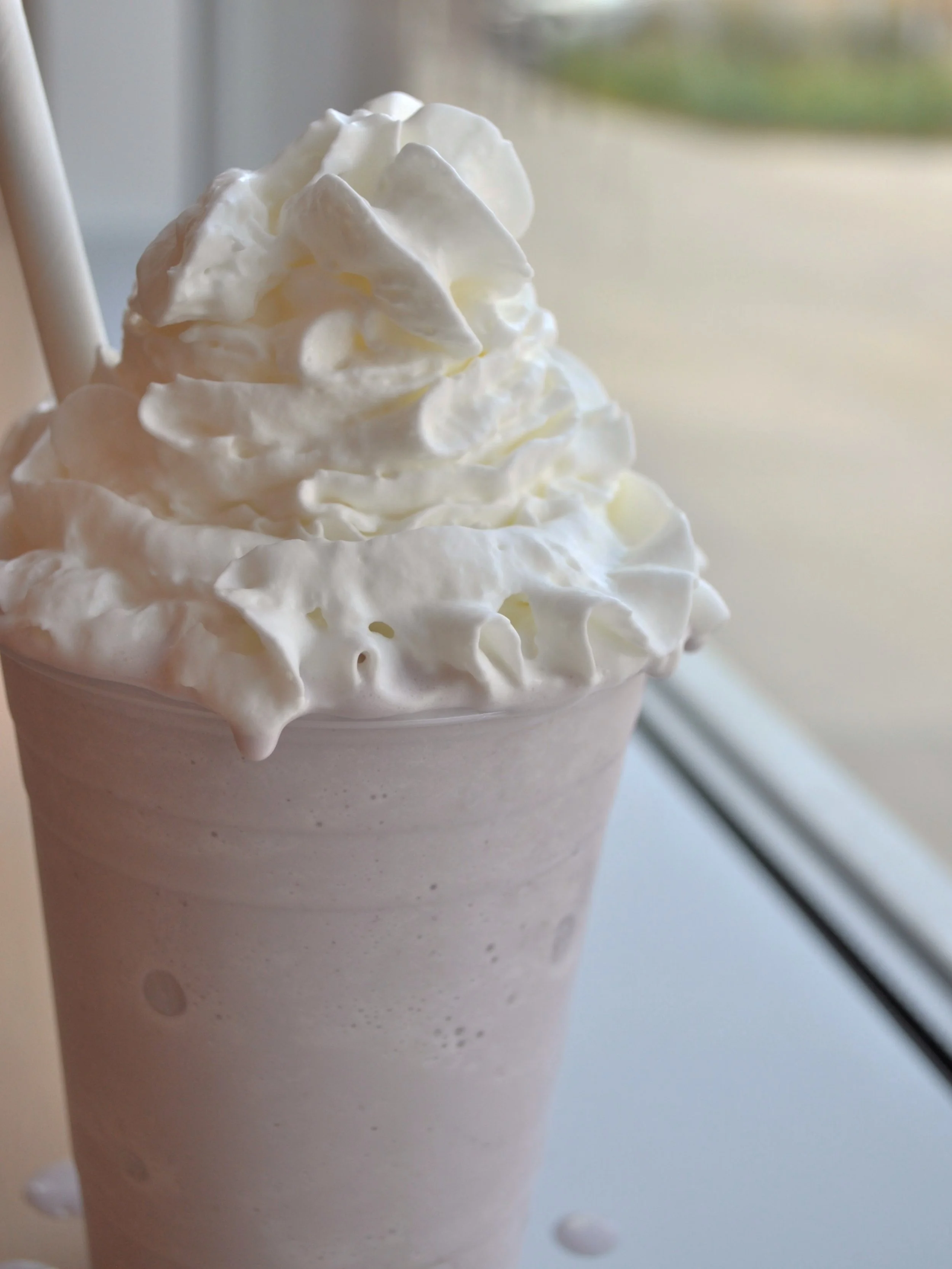 A creamy malted chocolate milkshake topped with drippy whipped cream placed near a window.