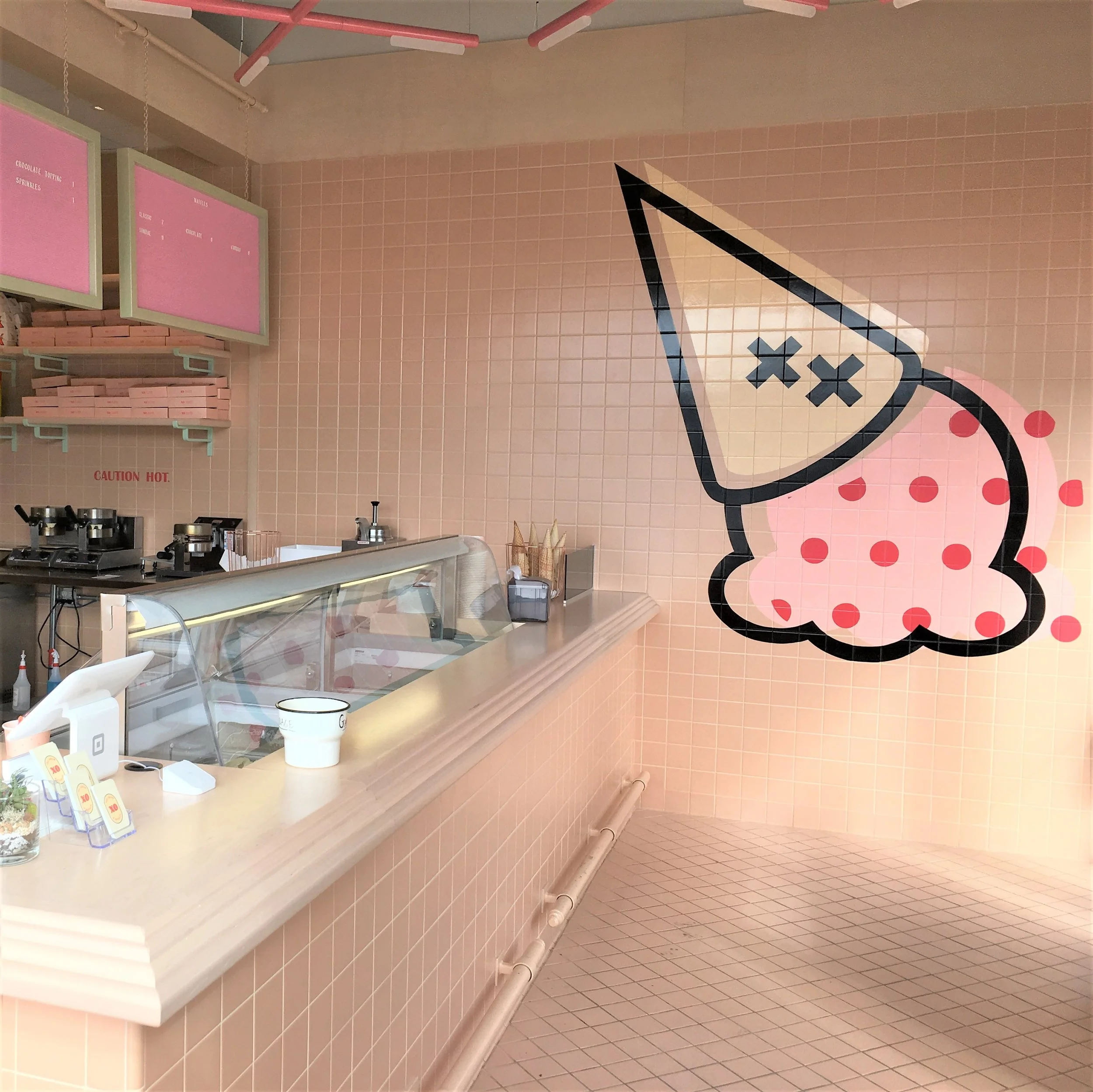 Interior of a pink ice cream or dessert shop with pink tiled walls, a large cartoonish ice cream cone mural on the wall, pink menu boards, and a counter with a clear display case and some equipment.