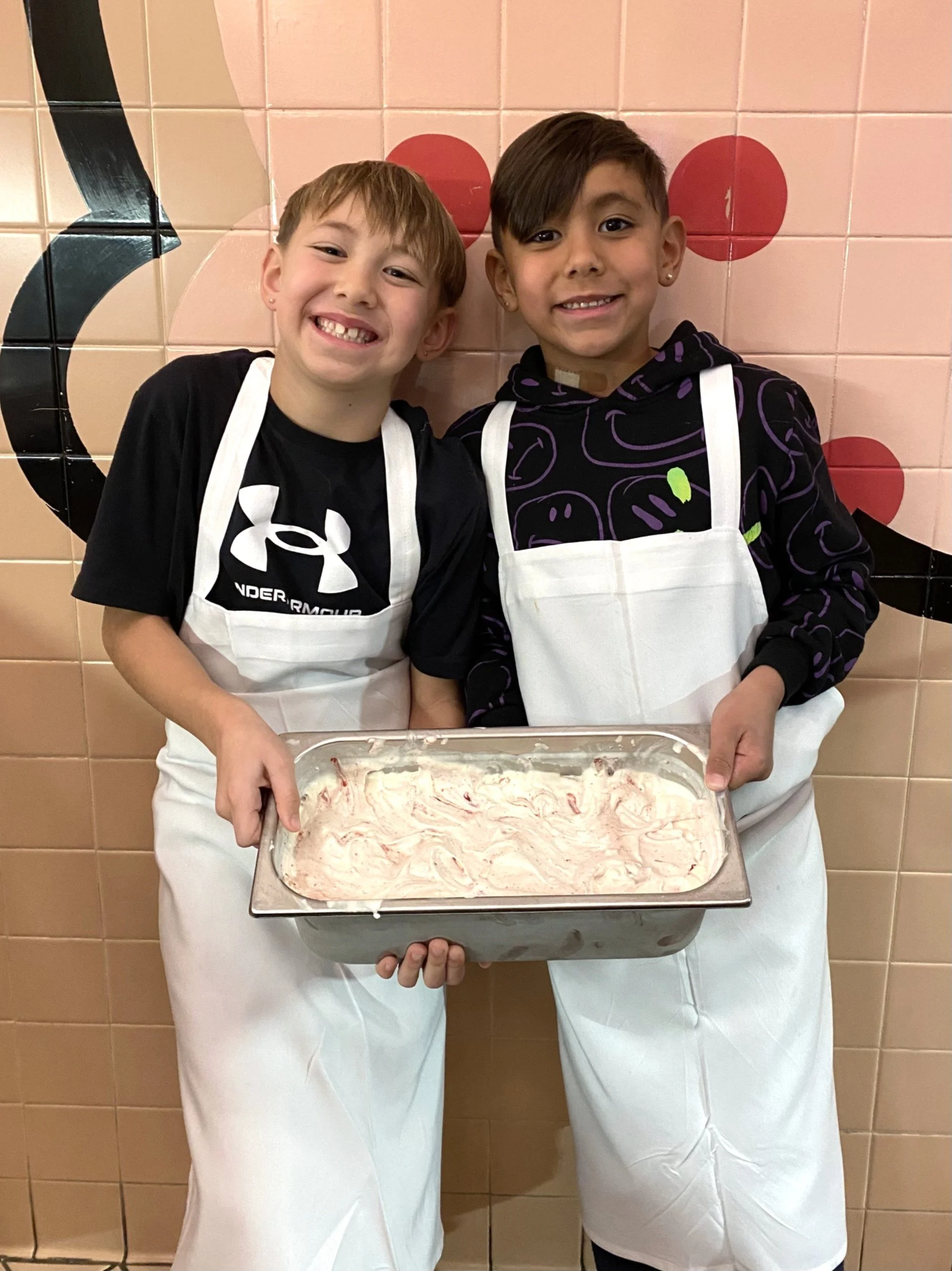Two children wearing white aprons, standing in front of a tiled wall with a black and red design, holding a batch of the ice cream they just made after winning the Back-to-School contest.