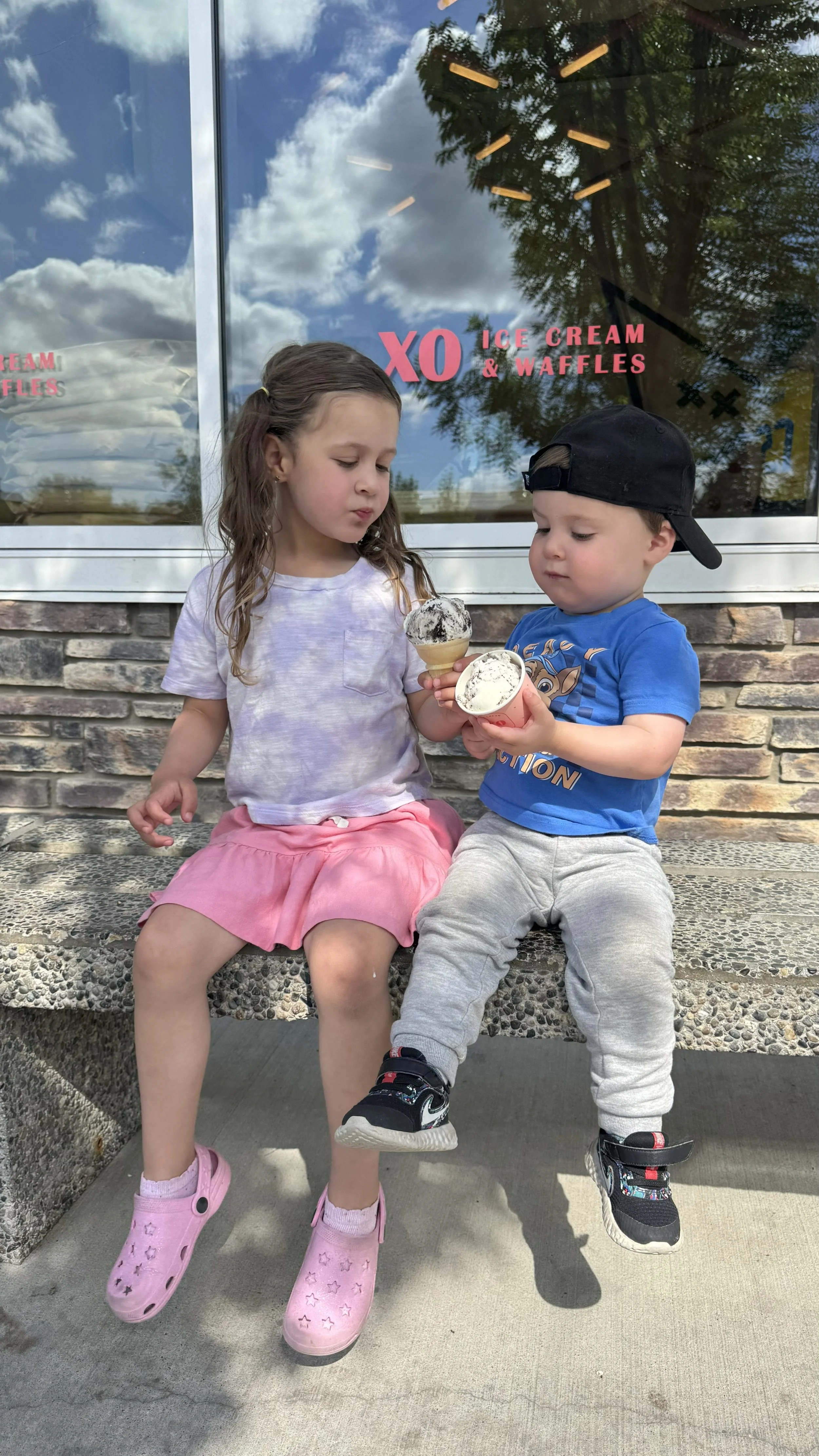 Two children, a girl and a boy, sit on a bench outside an ice cream shop. They are both holding cups of ice cream and are looking at each other. The girl is wearing a light-colored tie-dye shirt, pink skirt, and pink Crocs. The boy is wearing a blue shirt with a character design, gray sweatpants, a backwards black cap, and black sneakers with colorful accents. The shop window behind them displays the name 'XO Ice Cream & Waffles' and reflects a blue sky with clouds and nearby trees.