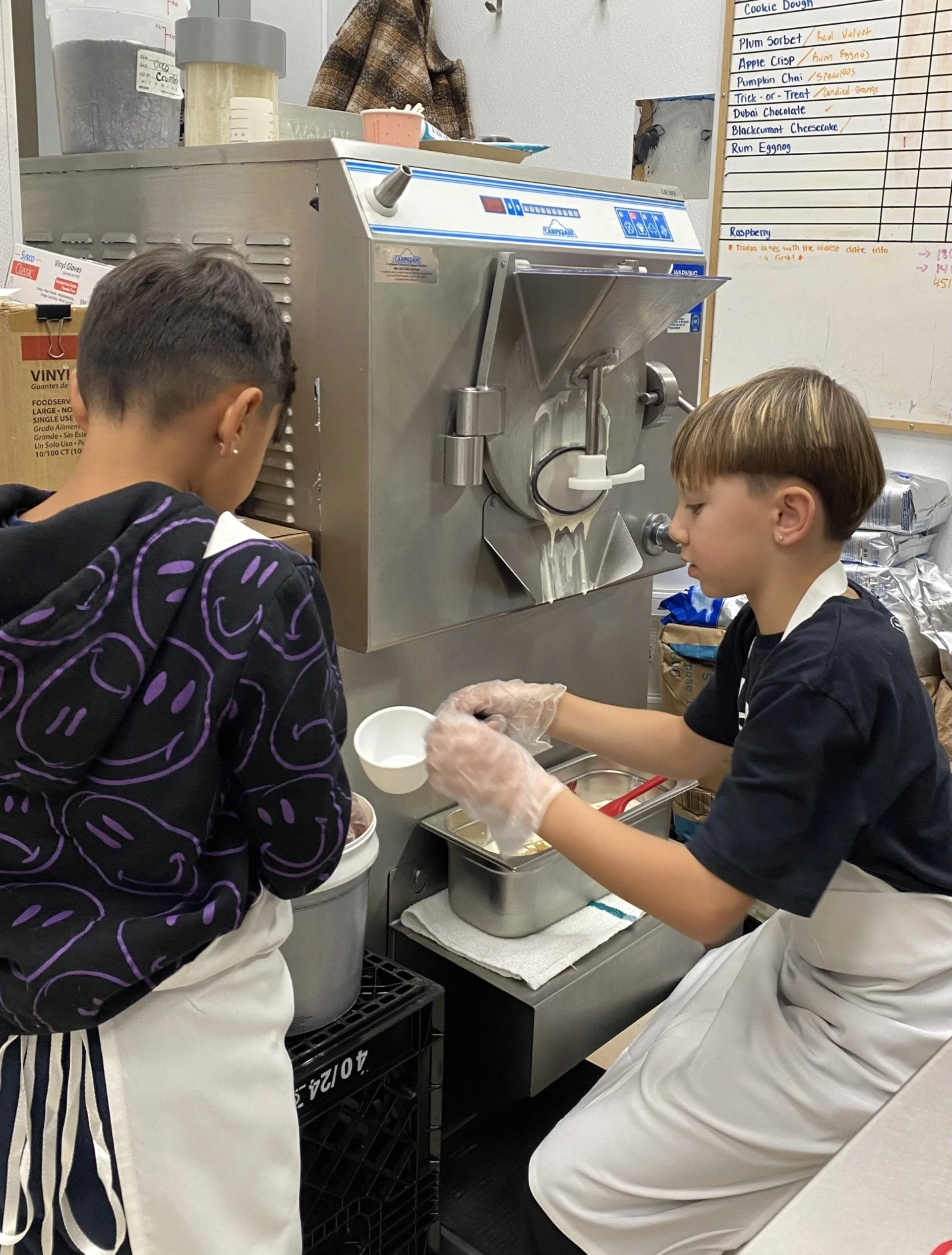 Two children wearing aprons working together in a kitchen: one with short dark hair, the other with short light brown hair. They are making a batch of their ice cream, after winning the Back-toSchool contest.