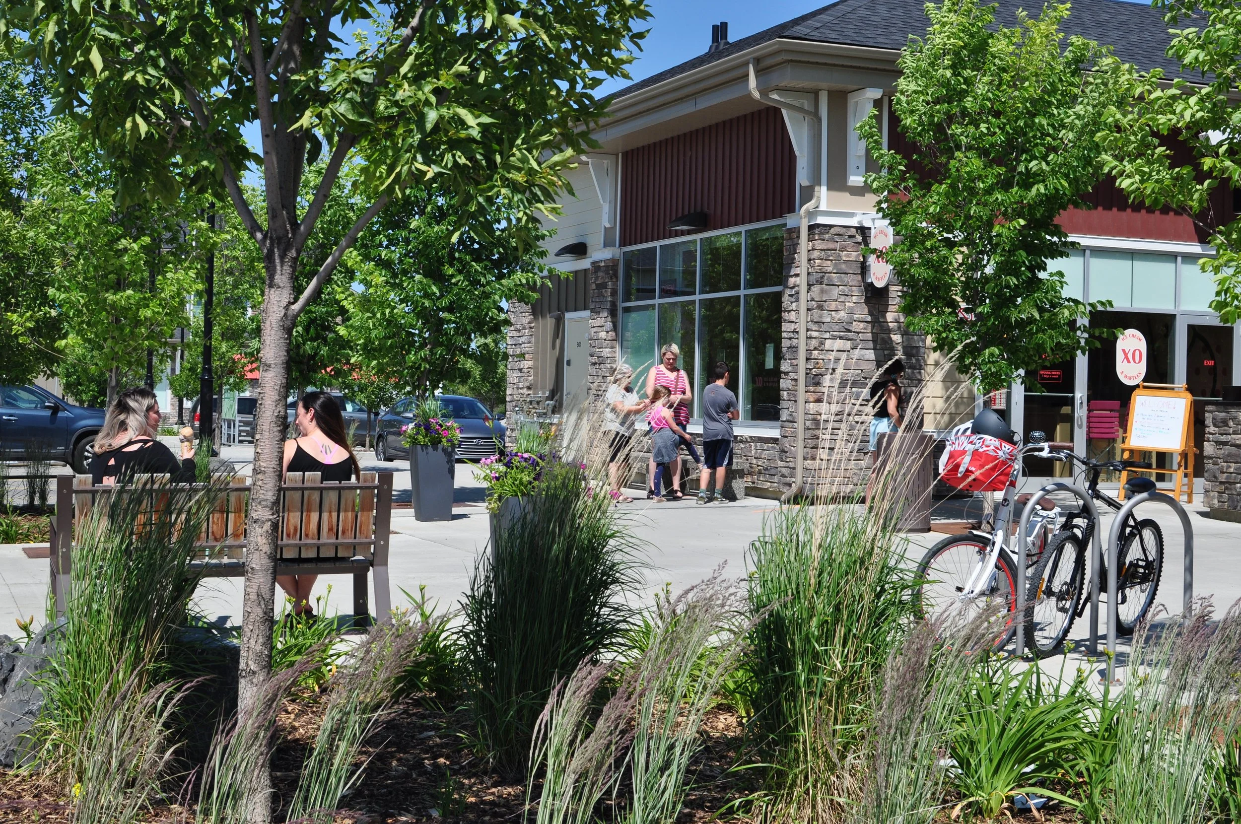 People sitting on benches and standing outside an ice cream and waffle shop called XO Ice Cream & Waffles on a sunny day, surrounded by trees and bicycles parked nearby.