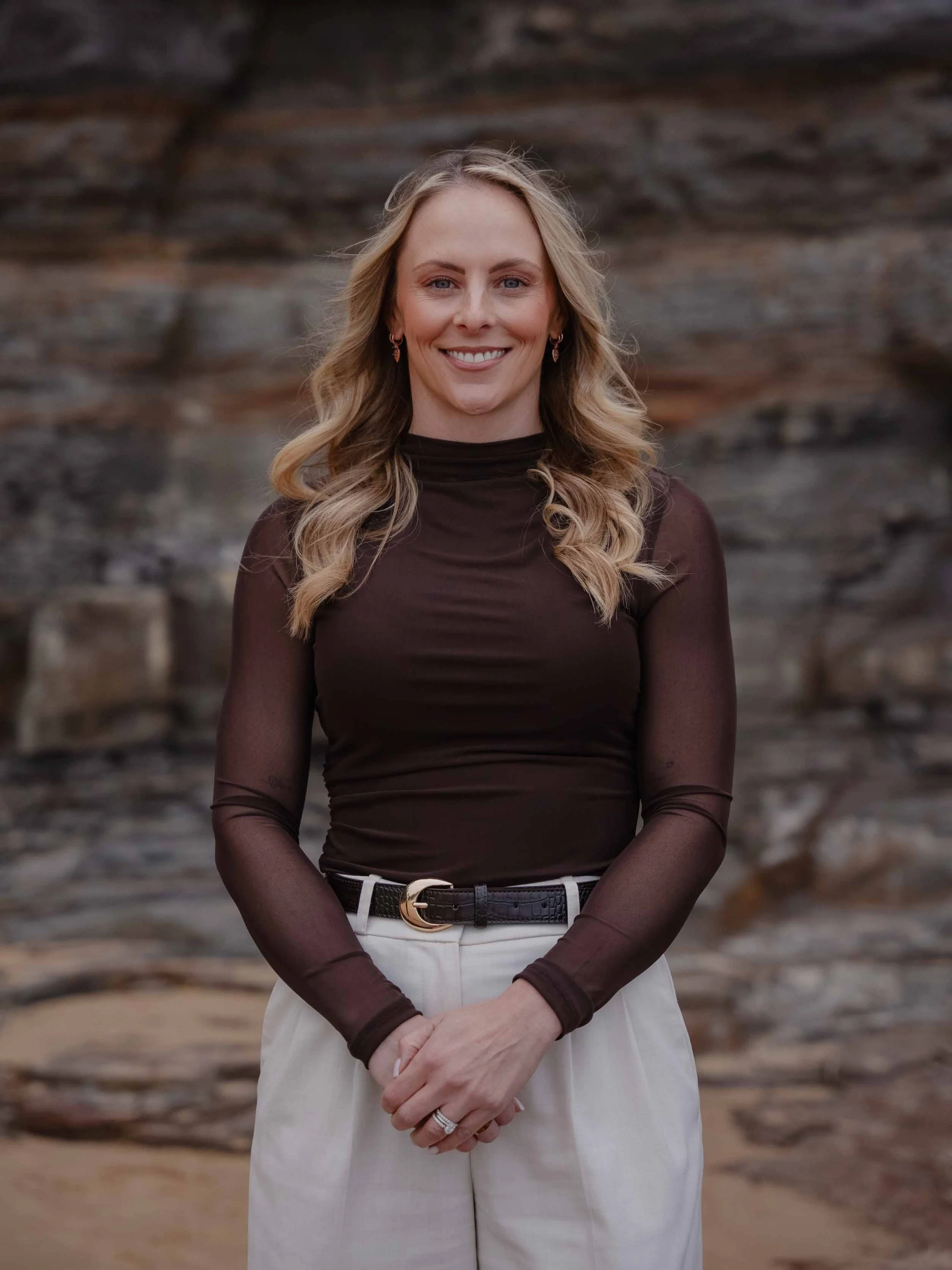 A woman with long blonde wavy hair smiling, wearing a dark sheer long-sleeved top and white pants with a belt, standing in front of a rocky background.