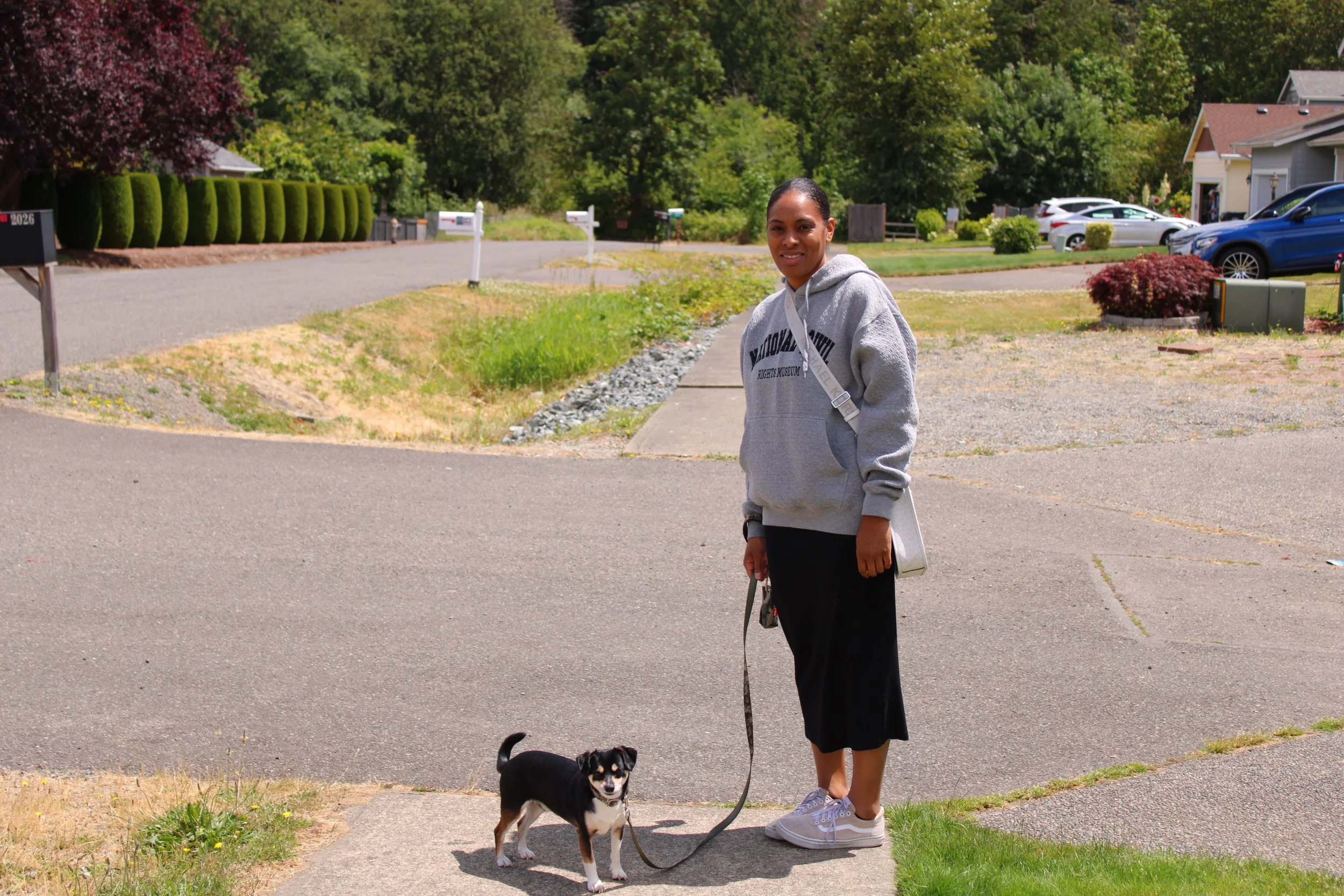 A woman in a gray hoodie and black pants holds a leash with a small black and white dog, standing on a sidewalk in a suburban neighborhood with greenery, houses, and parked cars in the background.