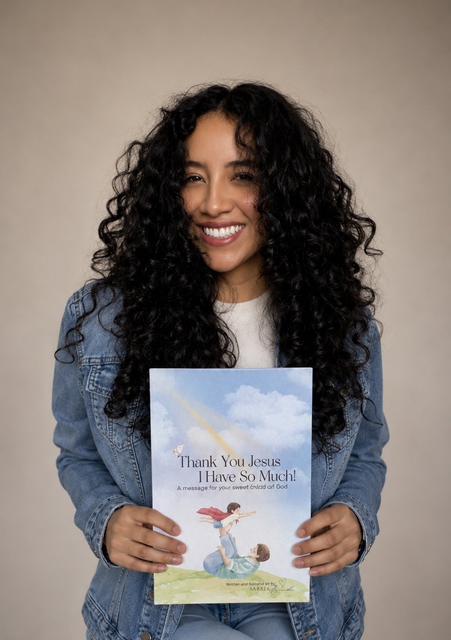 A woman with long, curly black hair smiling and holding a children's book titled "Thank You Jesus I Have So Much!" against a plain beige background. She is wearing a denim jacket over a white shirt.