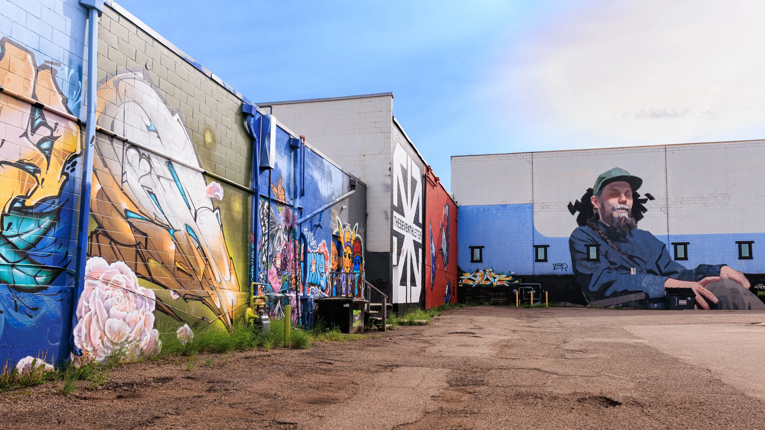 Colorful graffiti murals on the walls of a building, including a large portrait of a smiling man wearing a cap, in an outdoor urban setting.