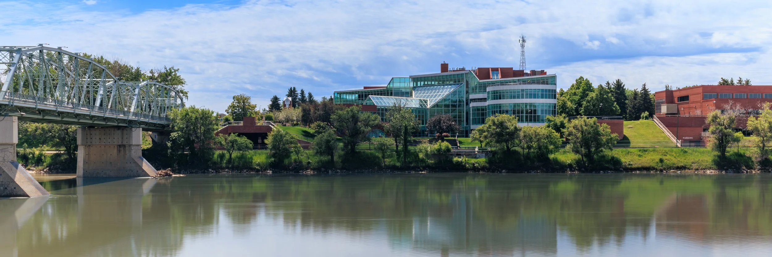 A landscape view of a river with a bridge on the left and modern glass buildings on the right, surrounded by green trees and grassy slopes under a partly cloudy sky.