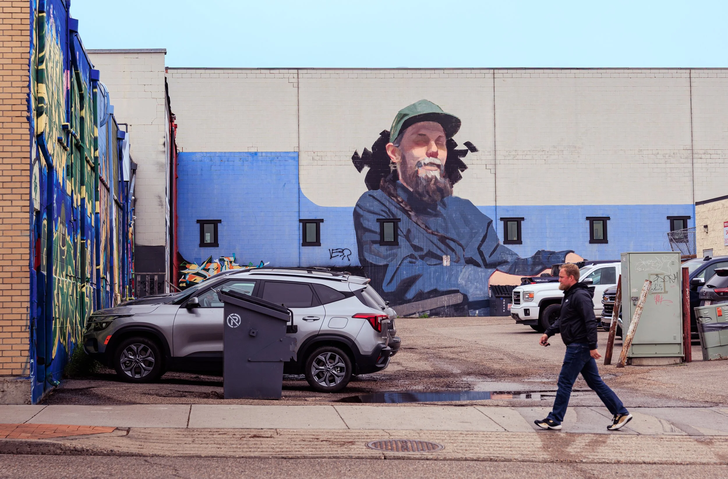 A mural of a man with a beard wearing a cap, painted on a large wall in an urban alleyway, with parked cars and a person walking in front.