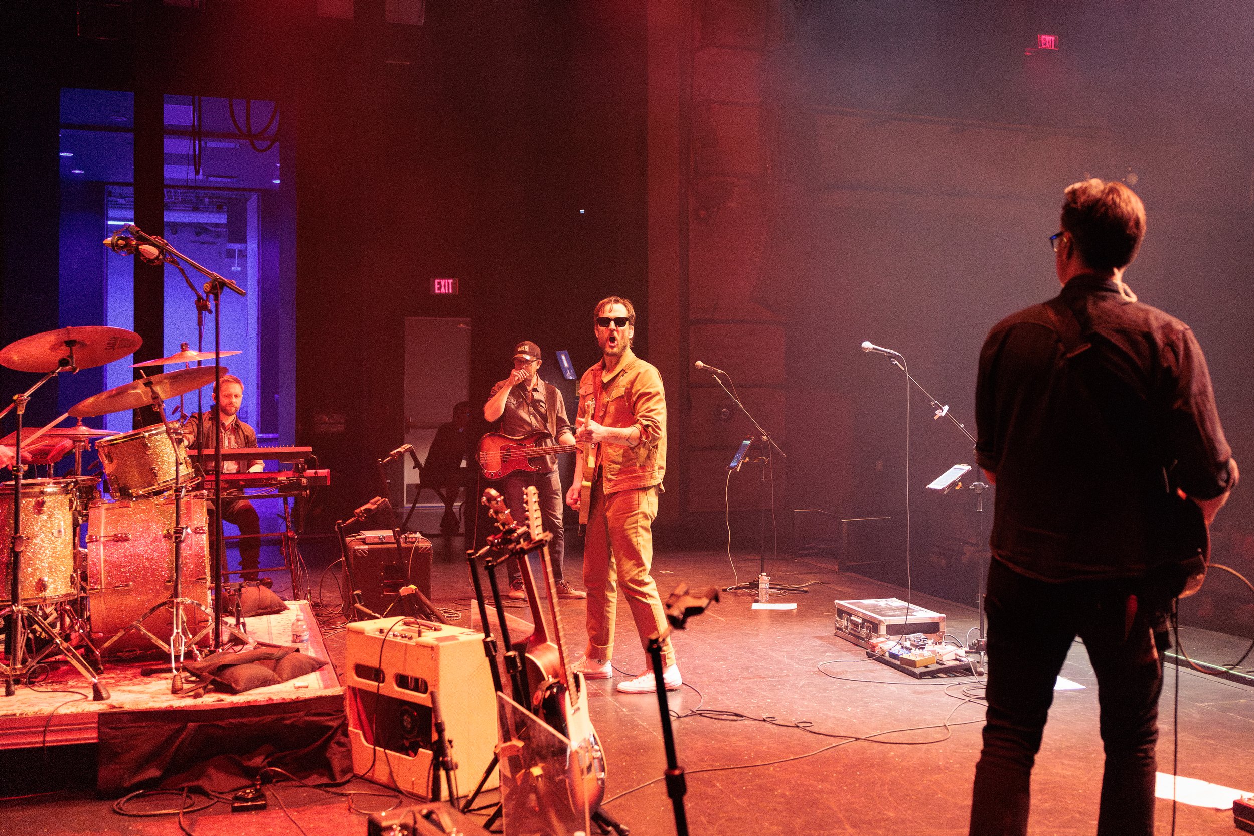 A band rehearsing on a stage with instruments, including drums, a guitar, and microphones. Several musicians are visible, one with sunglasses singing or speaking into a microphone, another playing guitar. The stage has equipment, and the background s