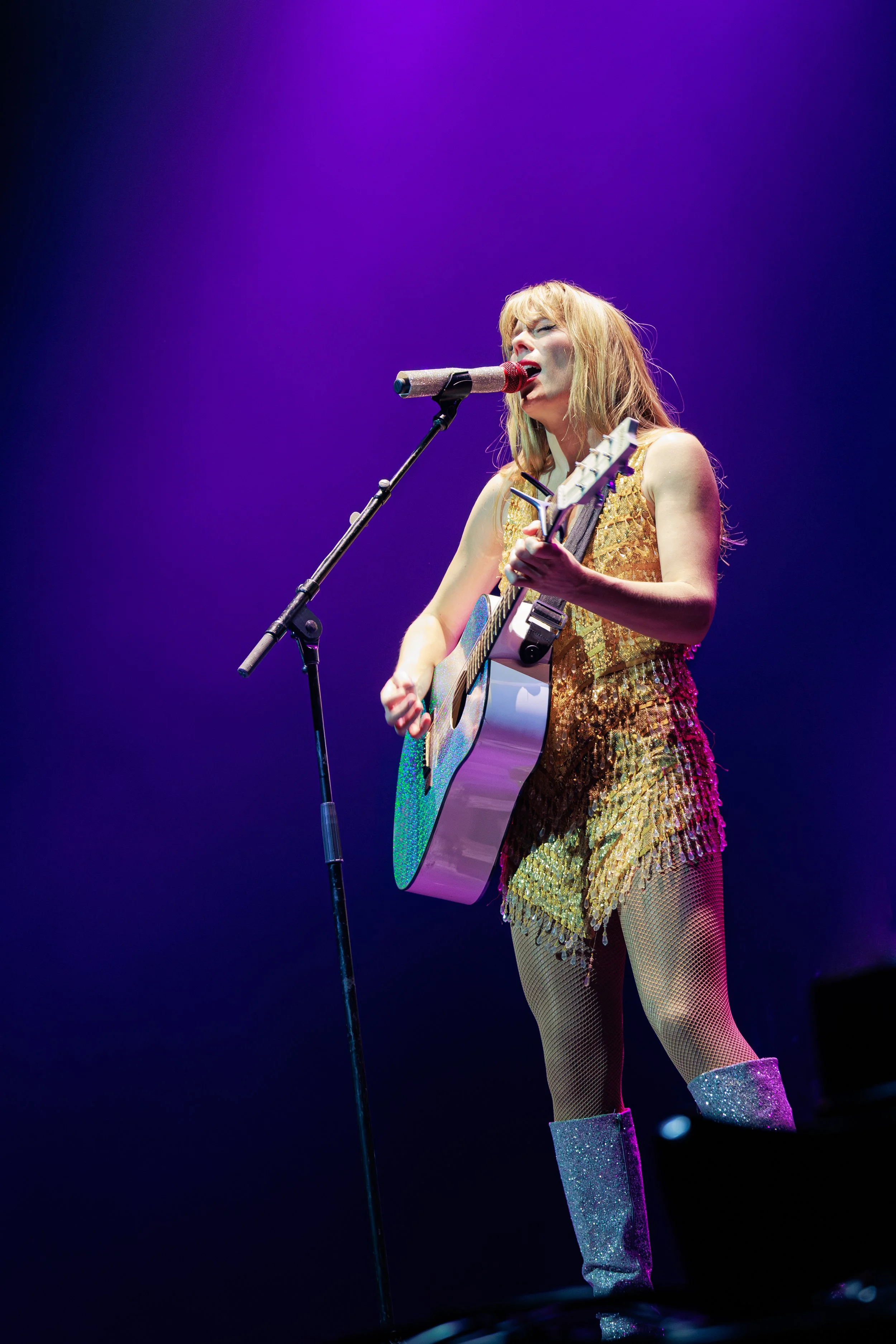 A female singer performing on stage with a guitar, wearing a shiny gold dress, fishnet stockings, and glittery boots, illuminated by colorful stage lights.