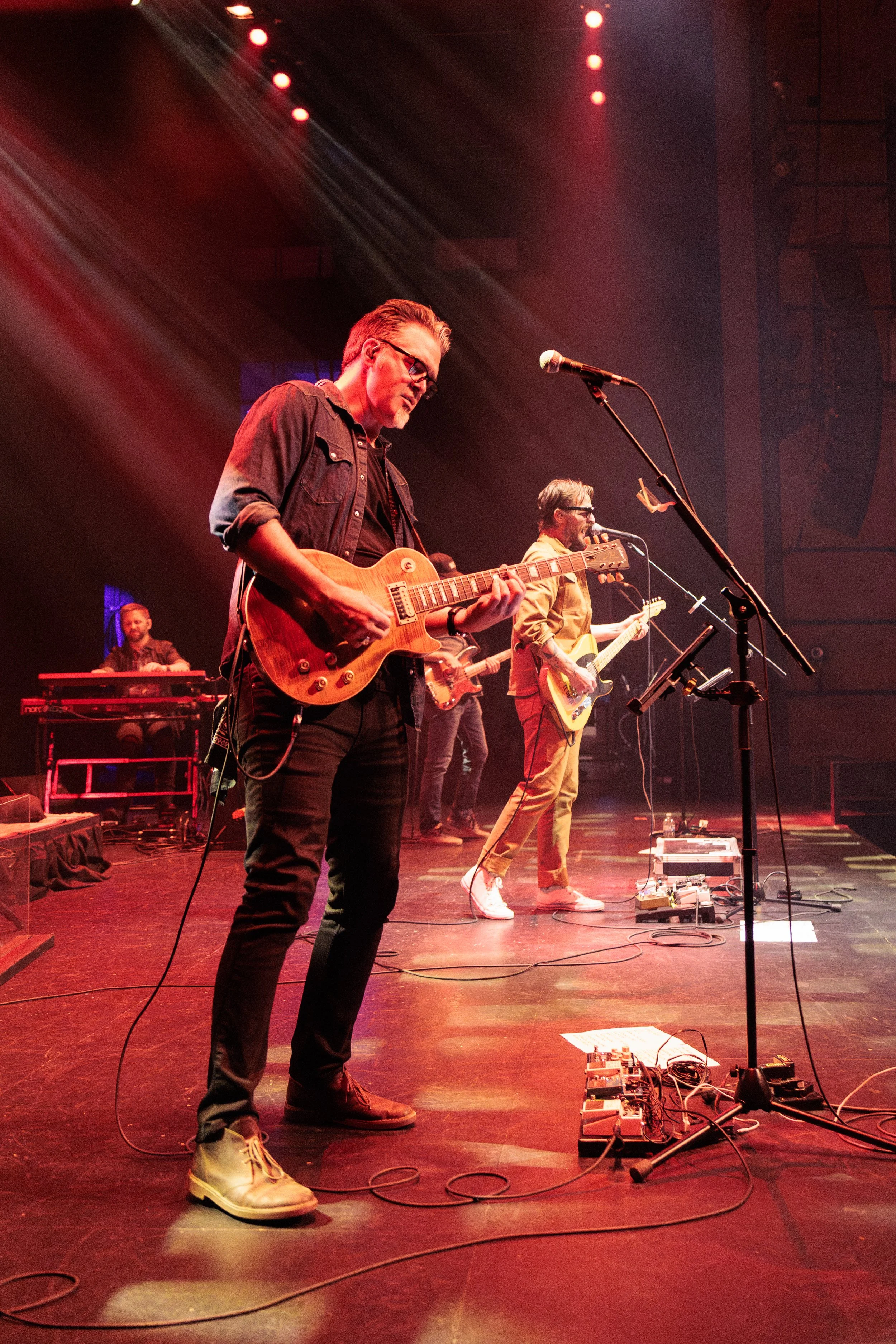 A band performs on stage at a concert, with one member playing an electric guitar in the foreground and two others playing guitars in the background. The stage is lit with red and white lights.