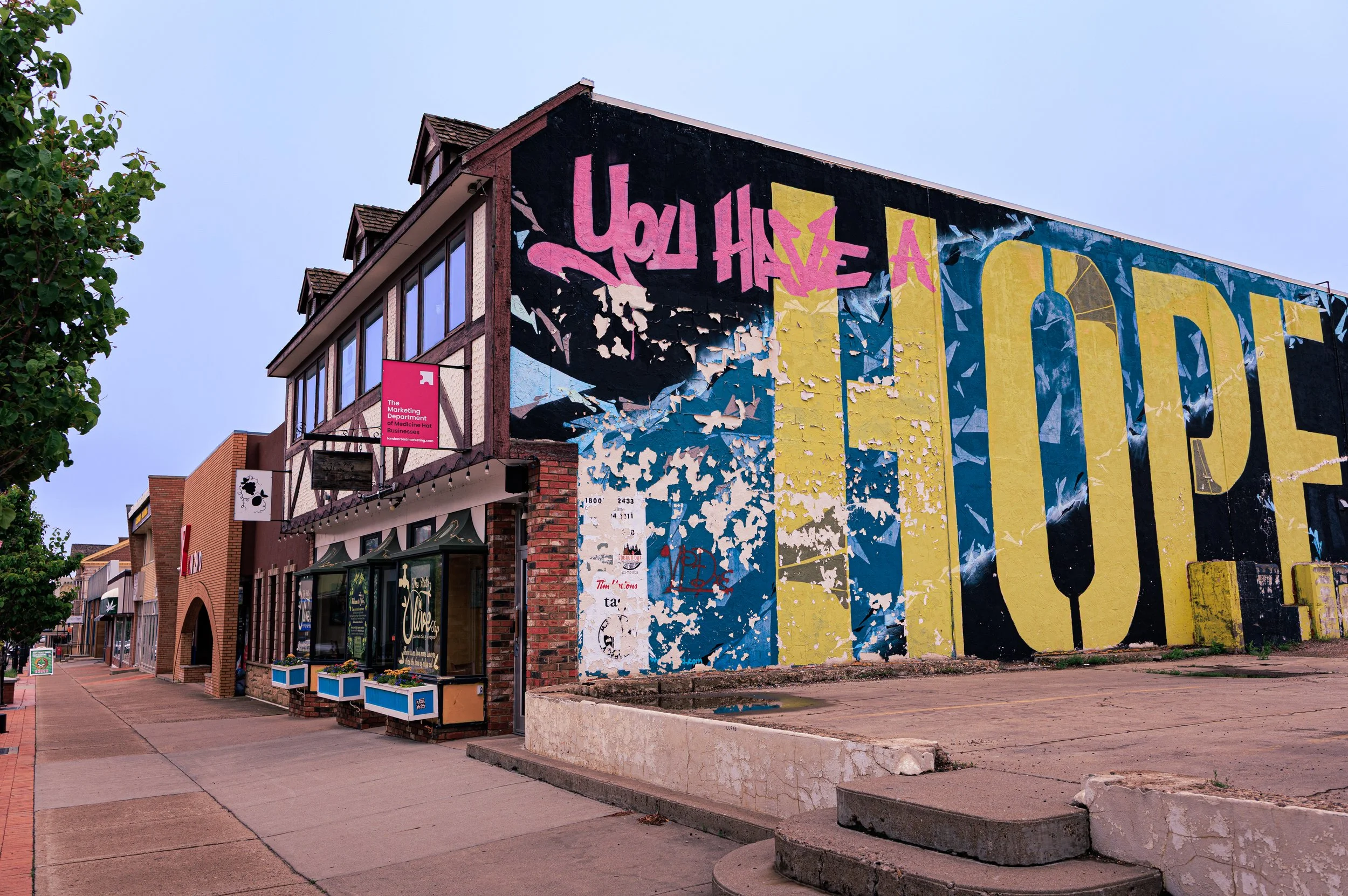 A street view with a large building featuring colorful graffiti art on its side wall, including the words 'You Have a' in pink. The atmosphere looks calm with storefronts and trees along the sidewalk.