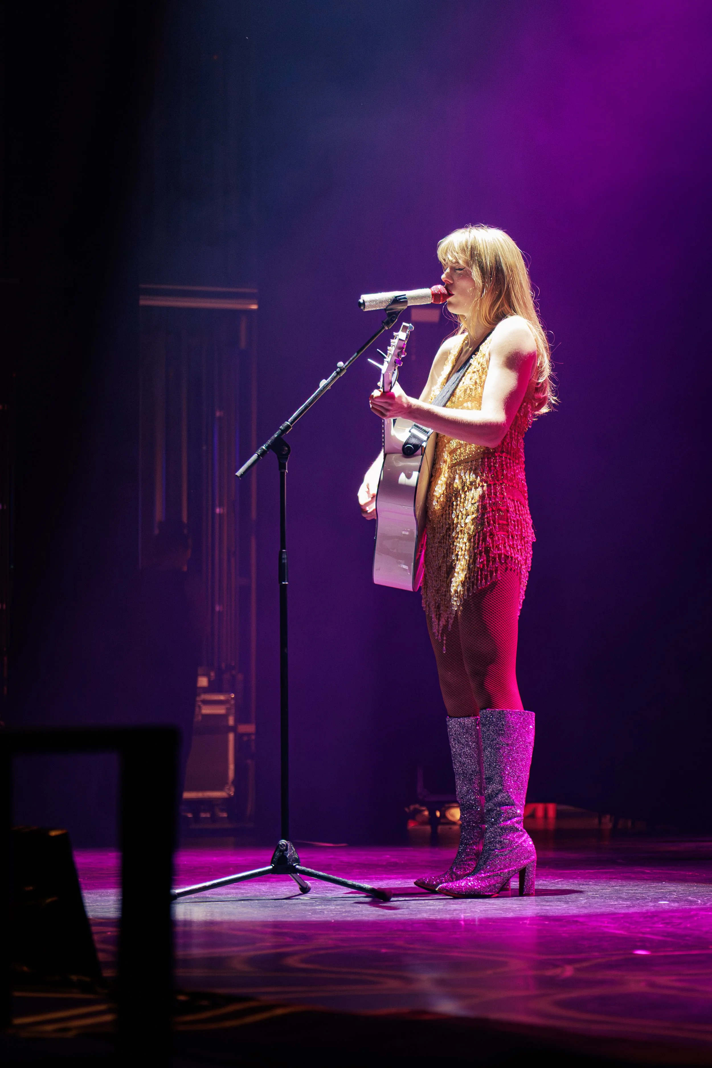 Female singer wearing a gold, sequined dress and glittery knee-high boots performs on stage with purple lighting, holding an acoustic guitar and singing into a microphone.
