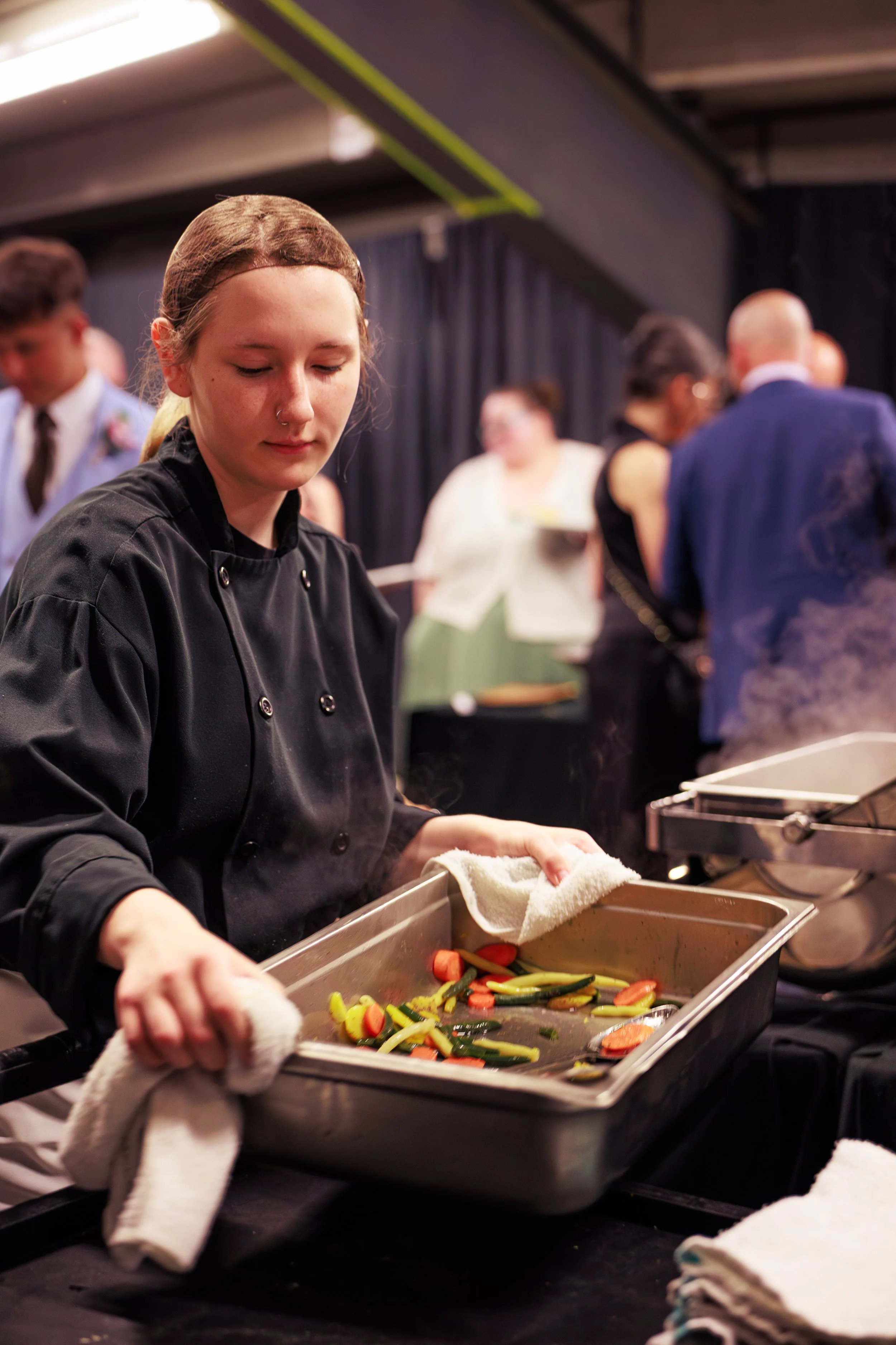 A young woman in a black chef's jacket preparing roasted vegetables, including carrots and green beans, on a baking tray in a busy kitchen or catering setting.