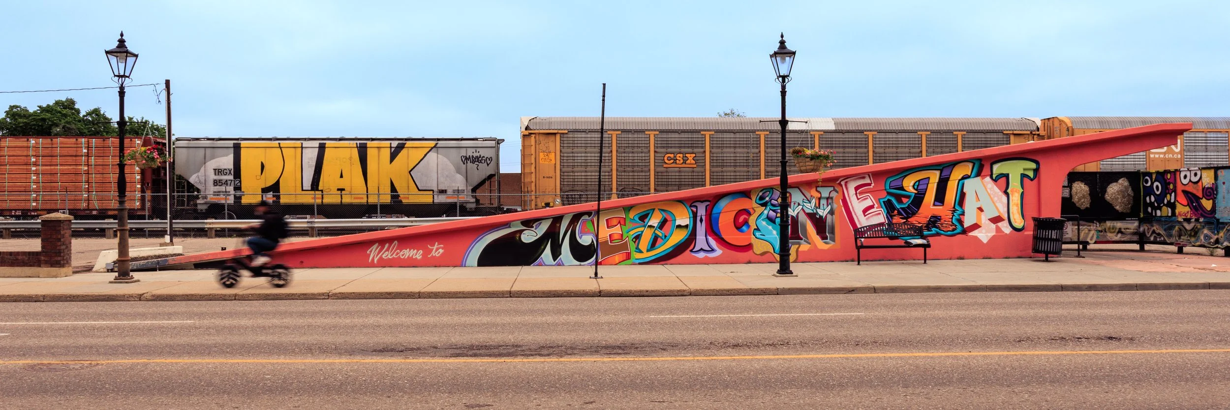 Colorful graffiti mural on a pink wall with benches and street lamps in front of a train yard.