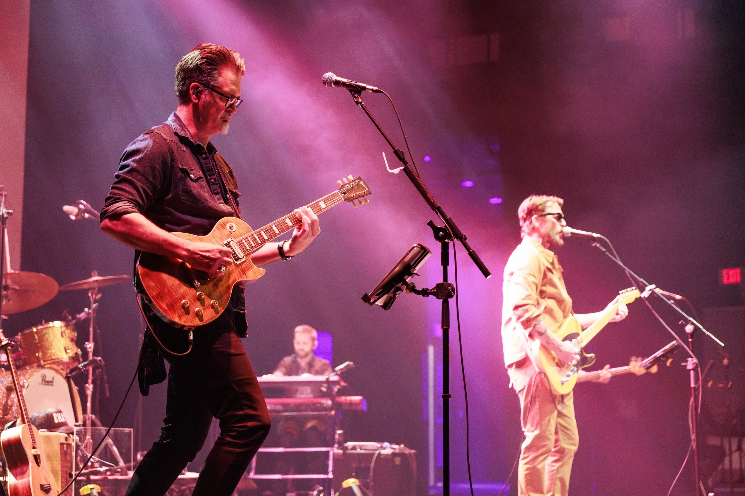 Musicians performing on stage, one playing an electric guitar and another singing into a microphone, with stage lights creating a vibrant atmosphere.