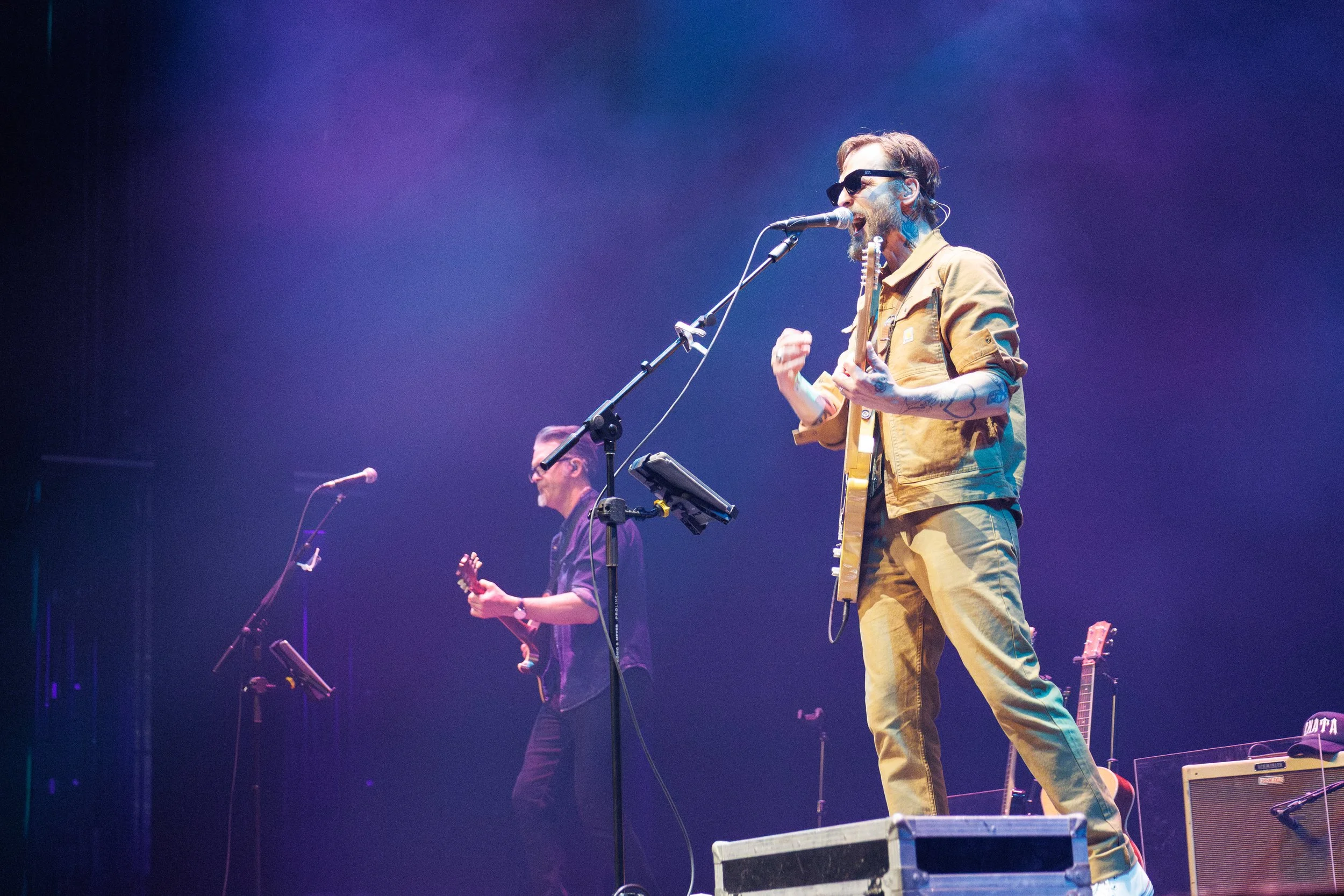 A male musician wearing sunglasses and a tan jacket, singing into a microphone and playing an electric guitar on stage with purple lighting. Another musician in the background is playing a guitar.