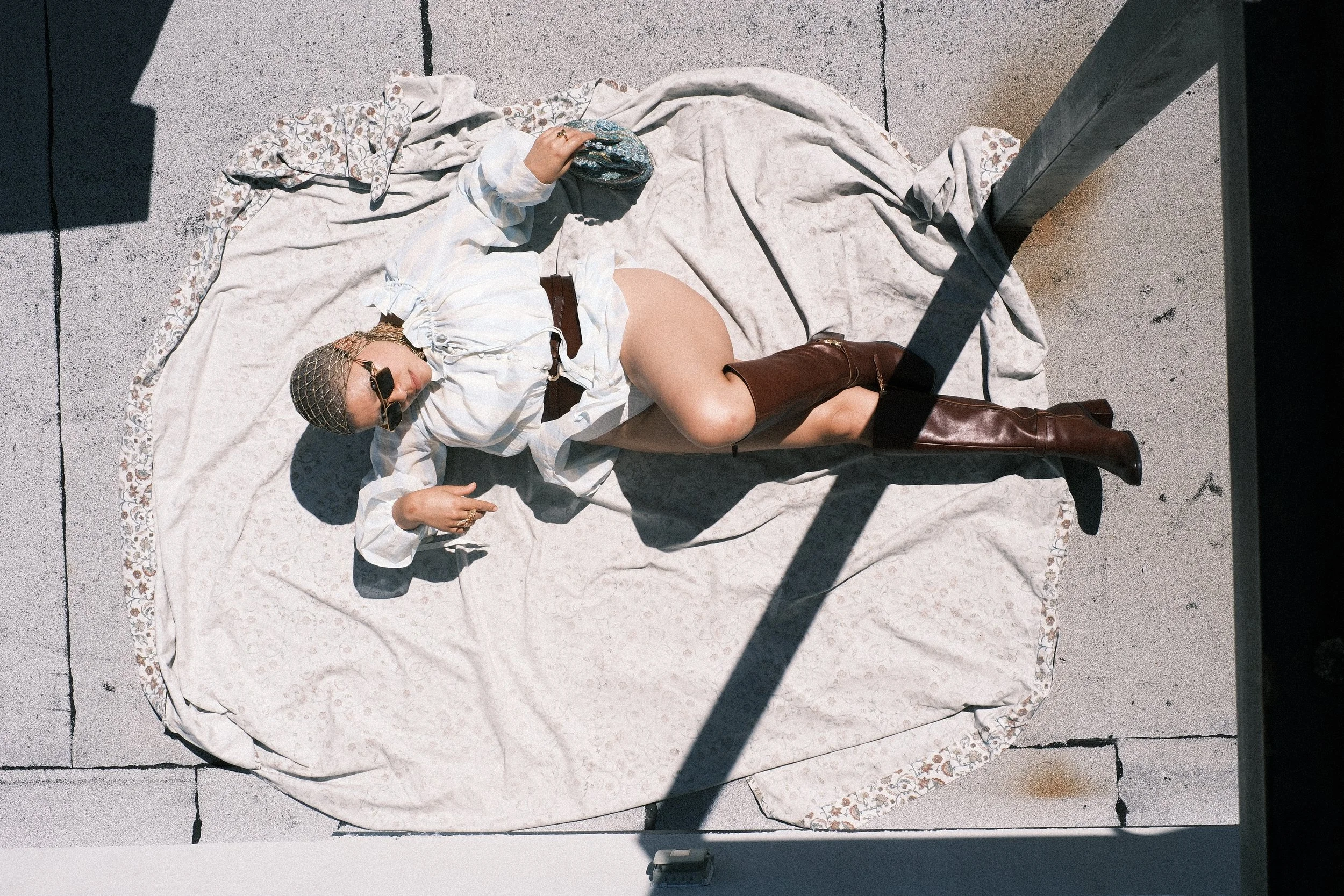 A woman lying on a patterned blanket on a concrete surface, holding a round, sparkly clutch bag, wearing sunglasses, a white blouse, and brown boots, with shadows cast across the scene.