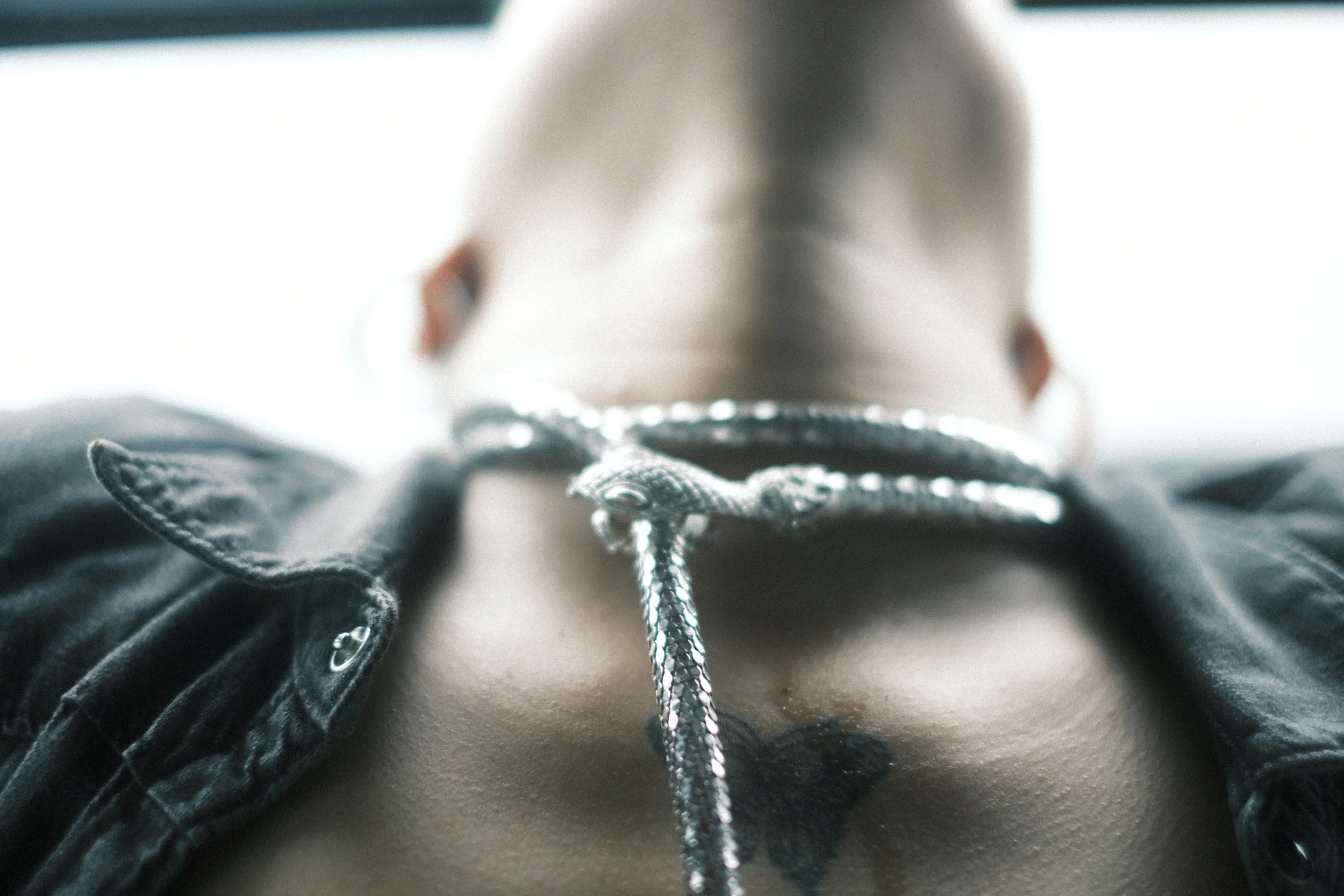 Close-up of a person lying out a window with a silver snake necklace on their chest, wearing a black denim jacket.