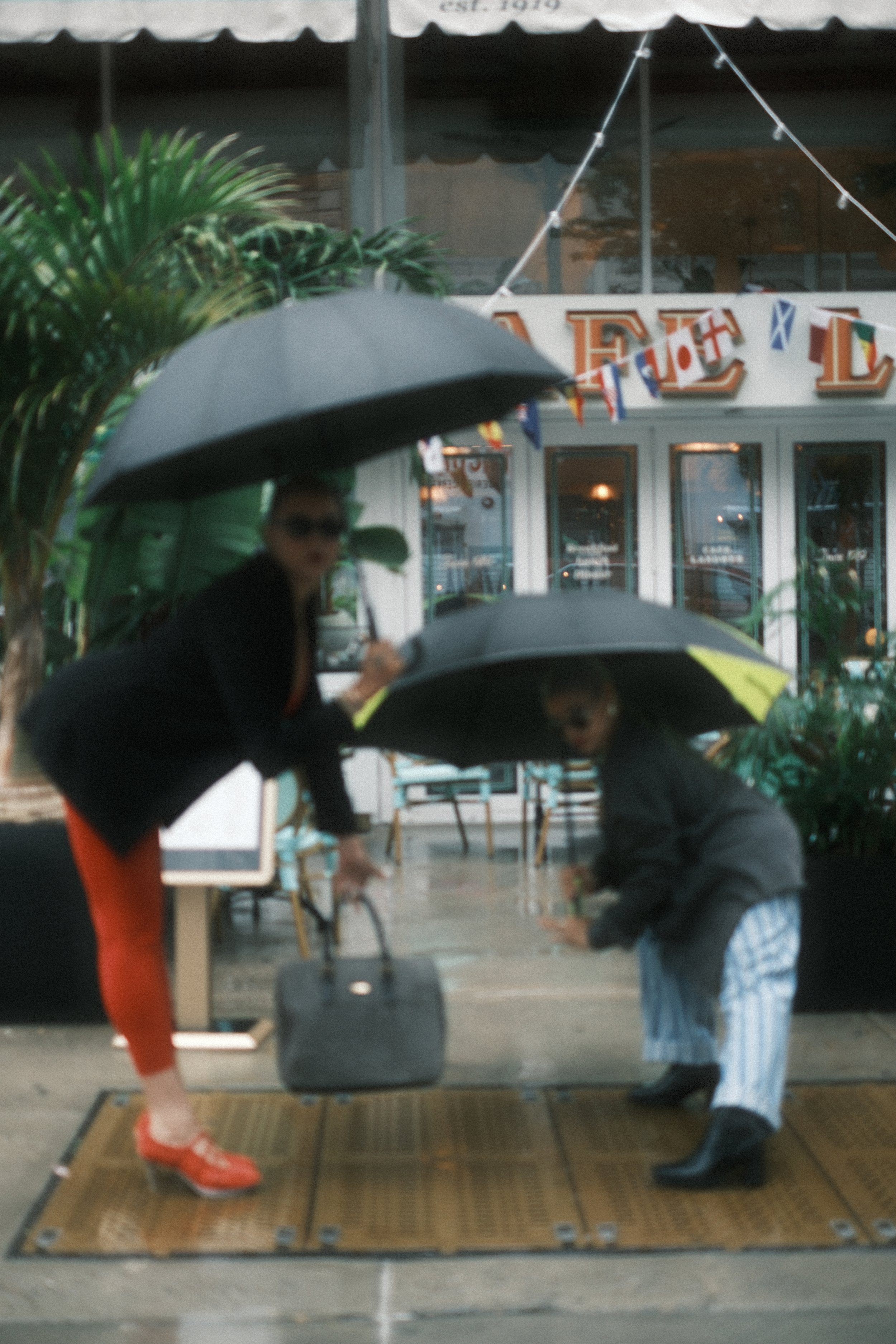 A woman with sunglasses, orange pants, and red shoes standing on a mat, handing a bag to a man in striped shorts and a dark jacket near the entrance of a restaurant or café, with a person being served at a table visible behind them.