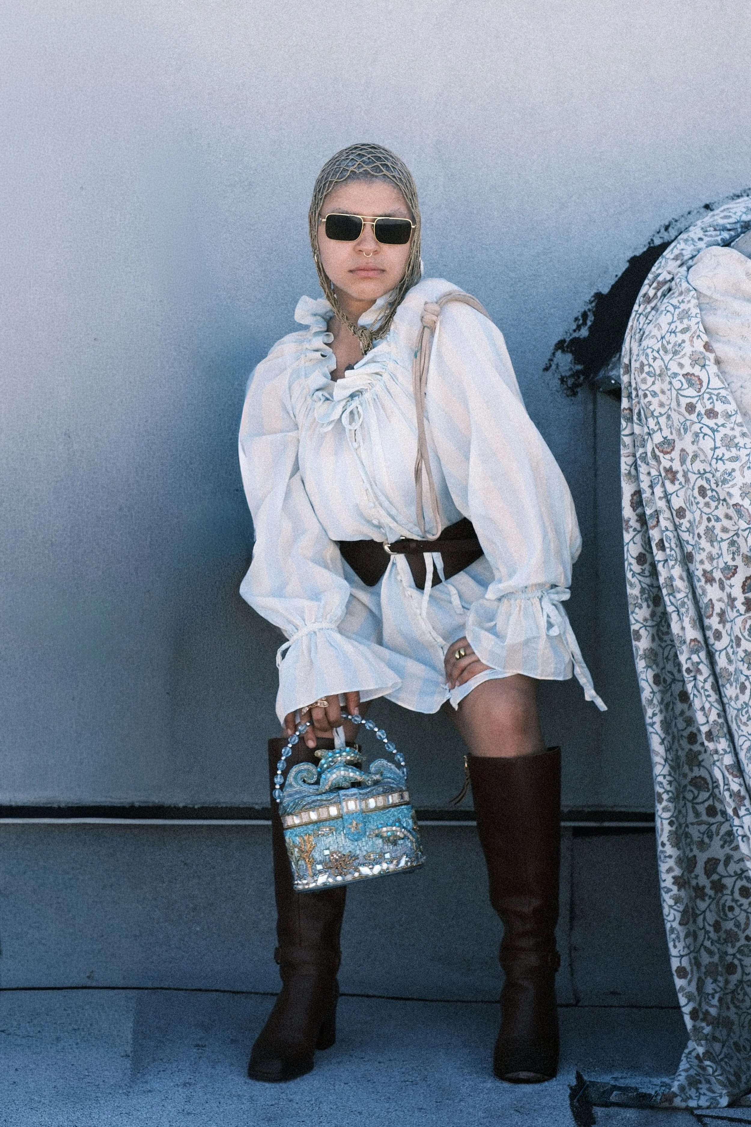 Person standing against a plain gray background, wearing a white ruffled blouse with puff sleeves, black thigh-high boots, and sunglasses. They are carrying a colorful, embroidered handbag and have a chain necklace. Part of a patterned curtain or fabric is visible on the right side of the image.
