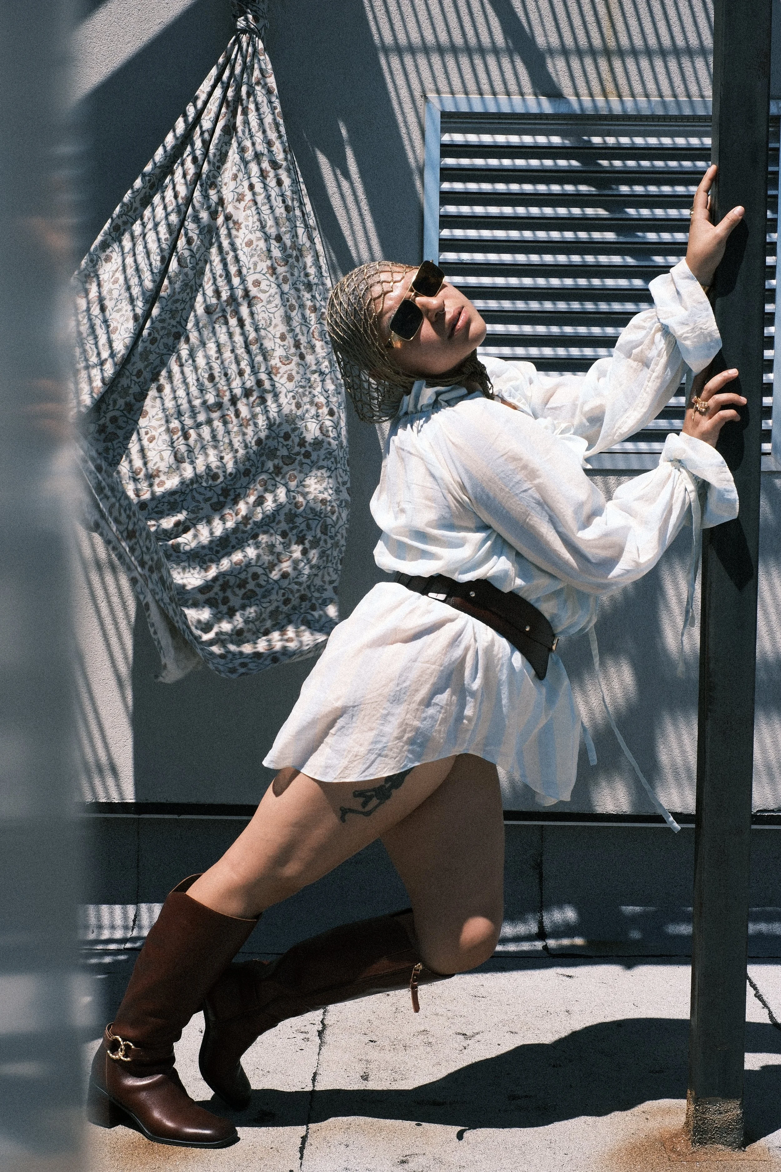 A person with wet hair and sunglasses lying against a gray wall with shadows, wearing a white oversized shirt and brown boots, with a tattoo visible on their thigh.
