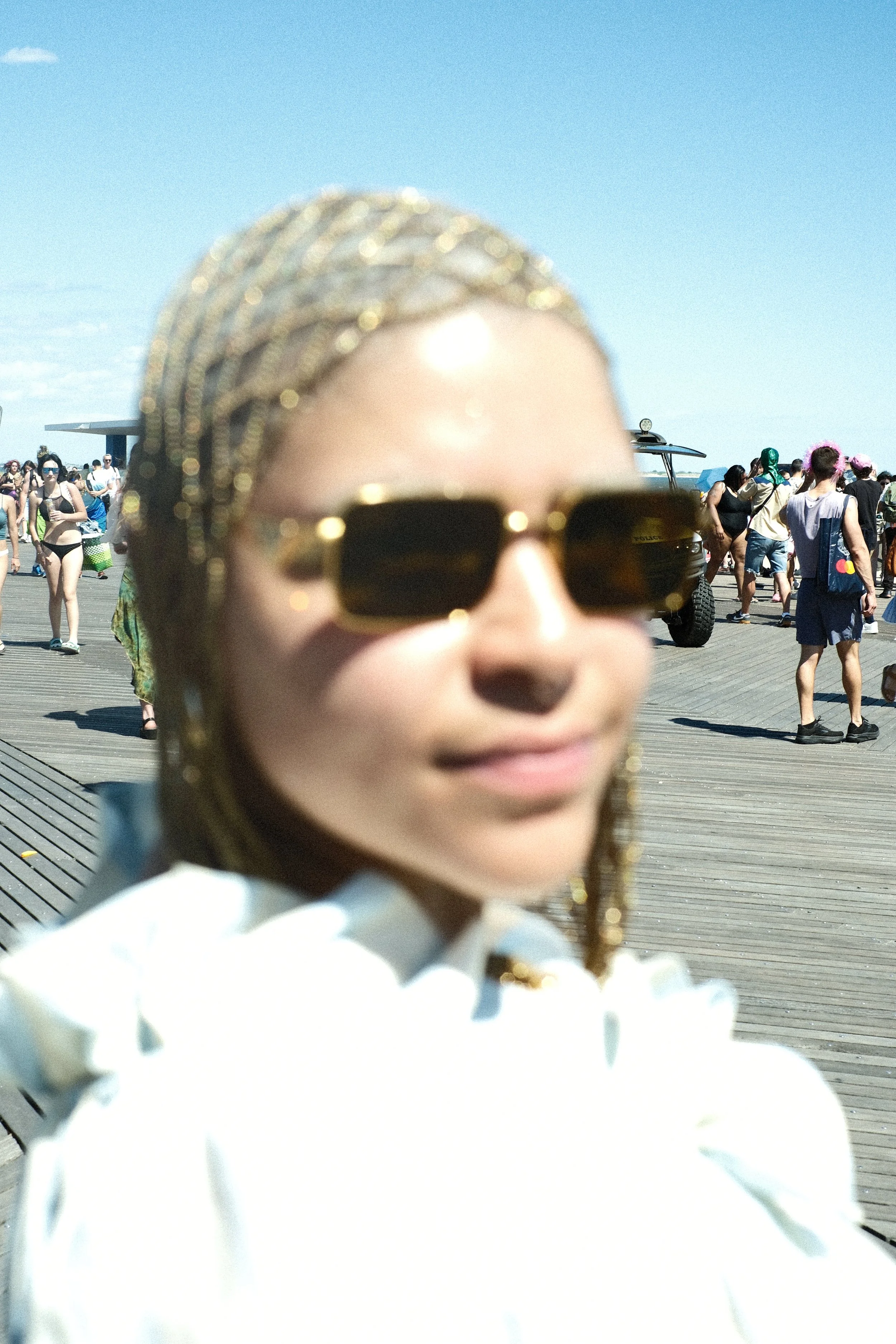 Close-up of a young girl wearing large sunglasses and gold earrings, with a background of people on a boardwalk near the beach, some in swimsuits and casual clothing, under a blue sky.