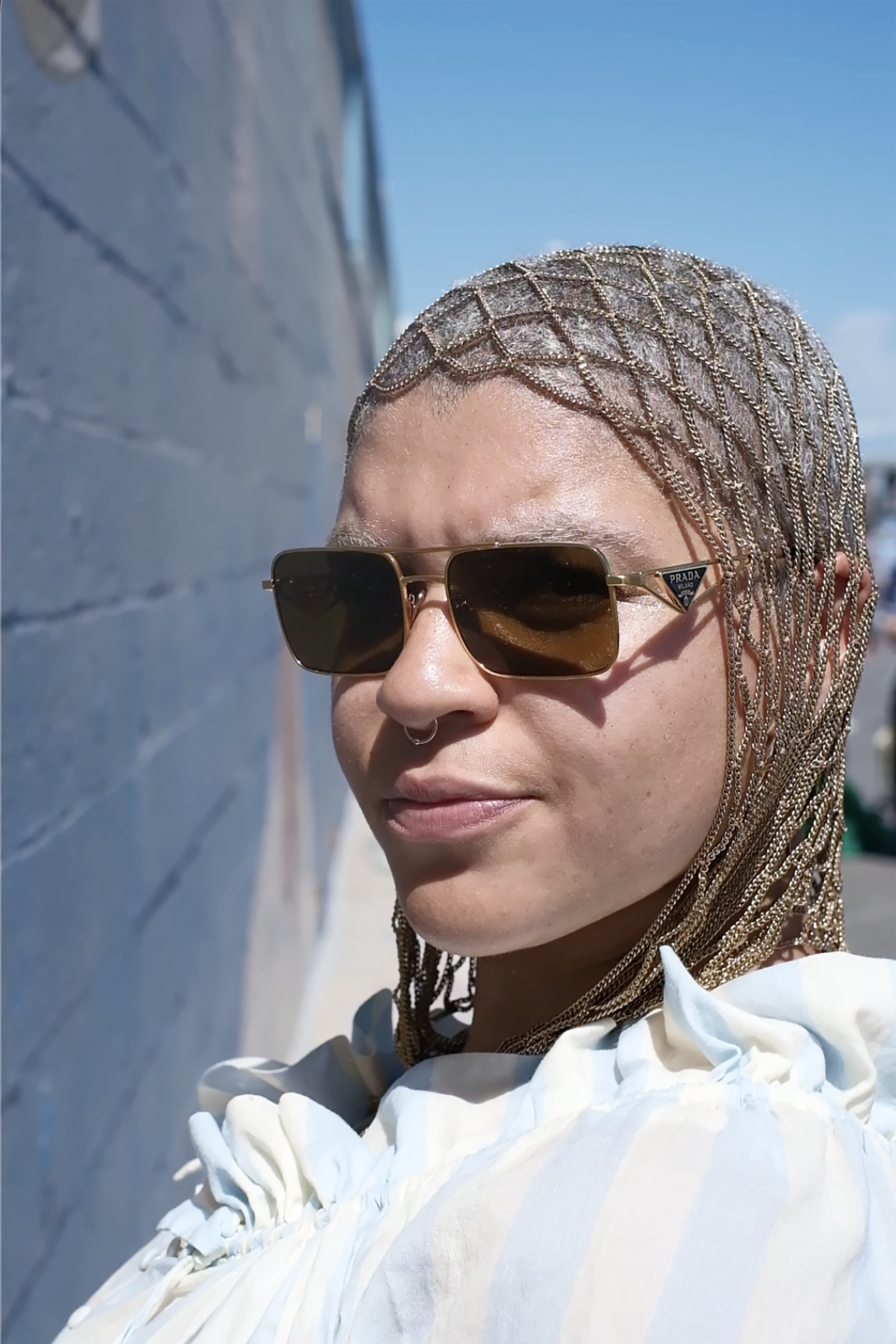 A person with short hair styled with a golden chain headpiece, wearing Prada sunglasses, a nose ring, and a white ruffled shirt, standing outdoors against a blue sky and a wall.