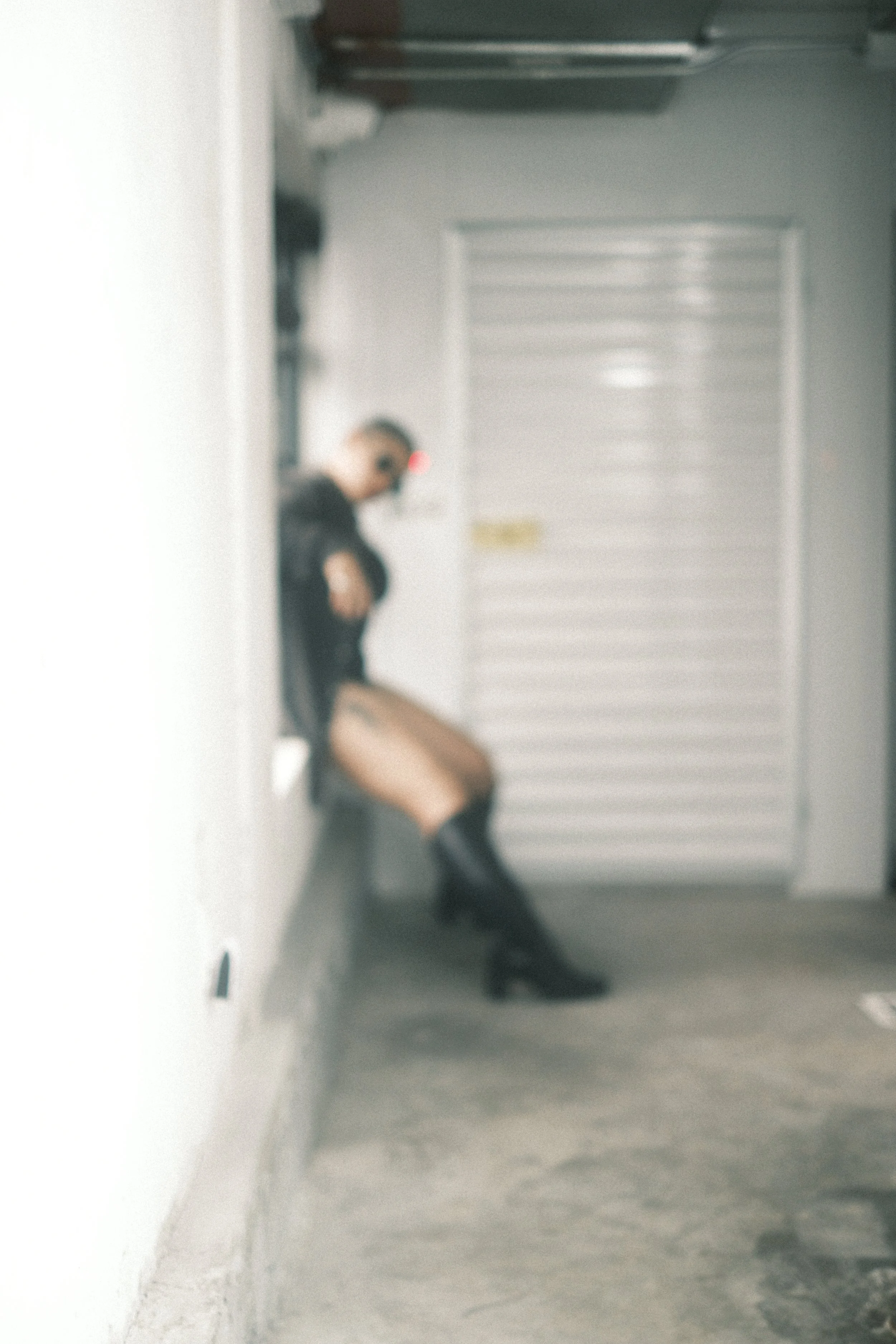 A person with blonde hair, wearing a black jacket, standing against a grey wall with a white door behind them. They are looking down and holding their face with one hand, while the other arm hangs loosely. The person is wearing black boots.