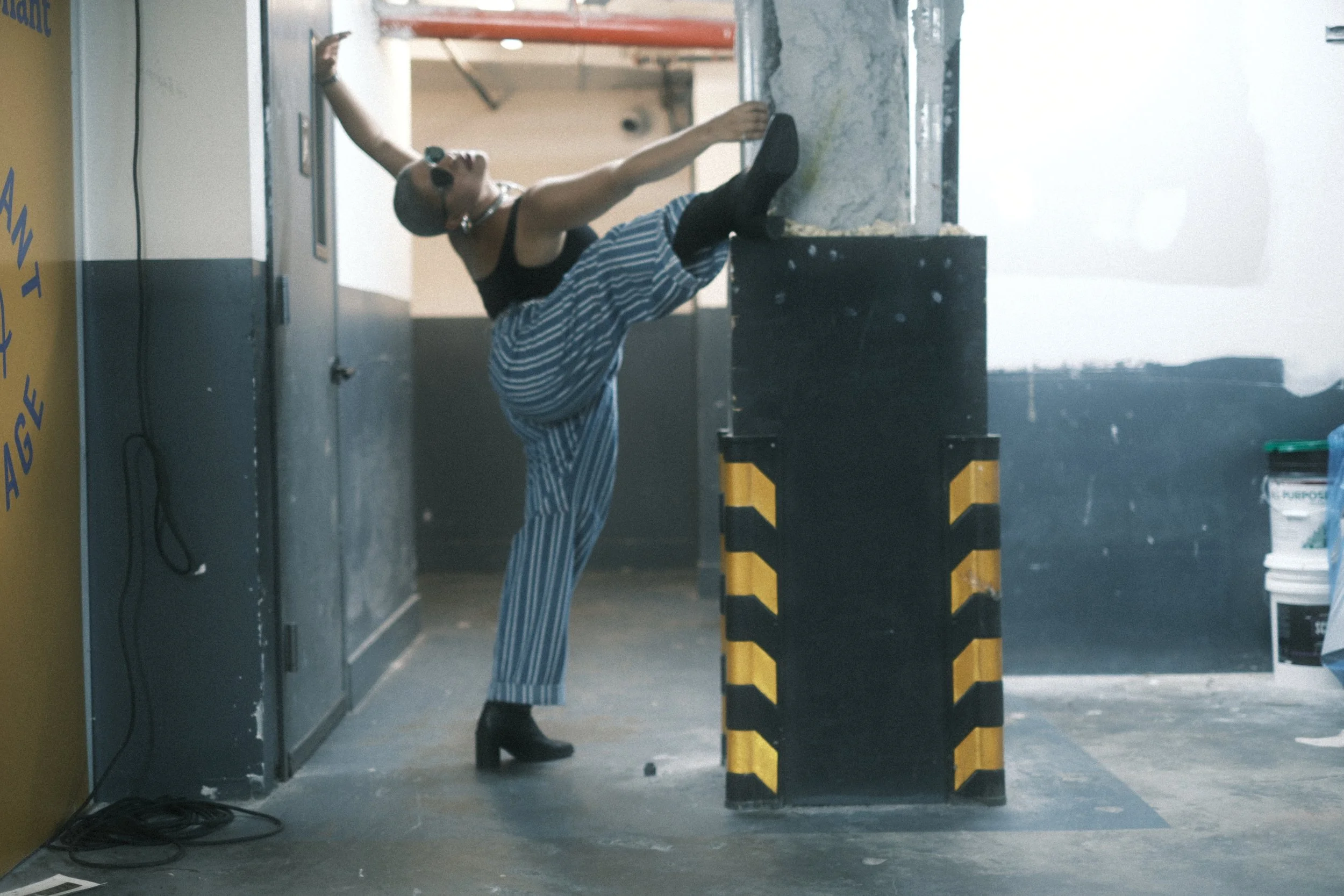 A woman standing on one leg, with her foot resting on a black and yellow striped barrier, poses in front of a mirror in a gym or fitness studio. She is wearing sunglasses, a black tank top, and striped pants, with her left arm raised above her head a