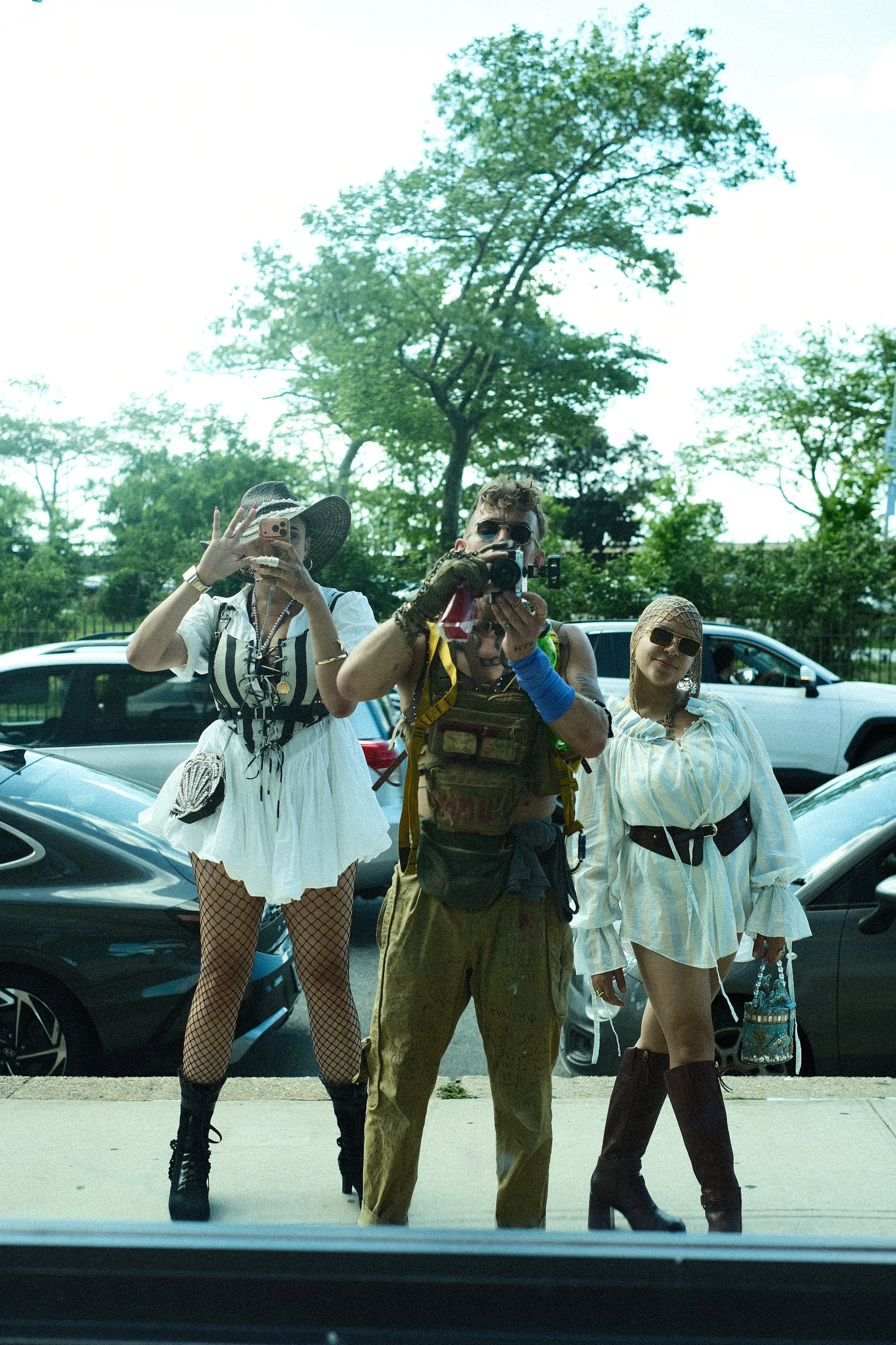 Three individuals dressed in fashionable and eclectic outfits pose in front of parked cars with trees in the background. The person on the left wears a white dress, fishnet stockings, black boots, and a large hat, and is taking a photo with their pho