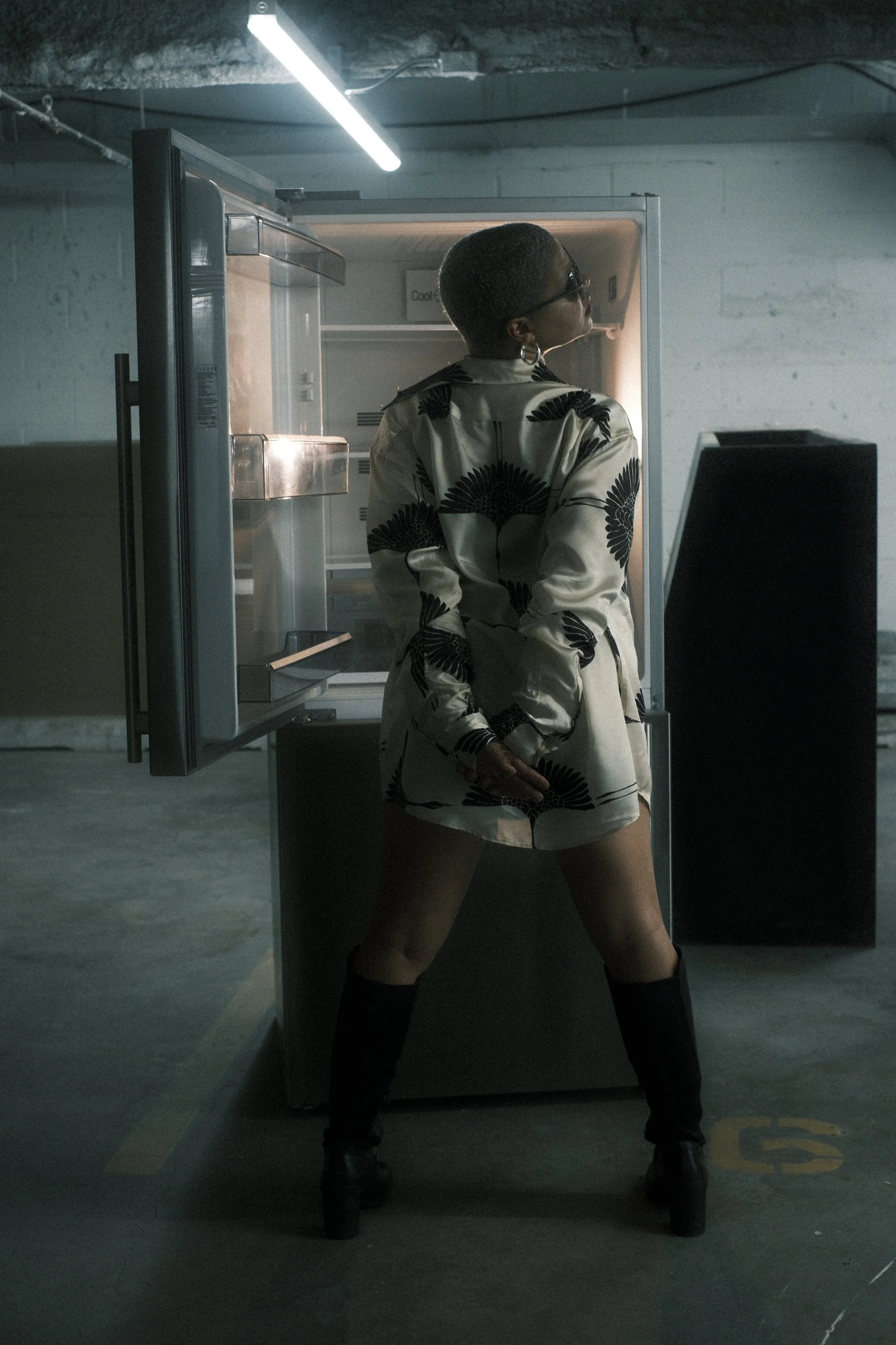 A woman with short dark hair and sunglasses standing in front of an open refrigerator in a parking garage. She is wearing a cream-colored jacket with black floral patterns, black knee-high boots, and shorts. The parking garage has dim lighting, with 