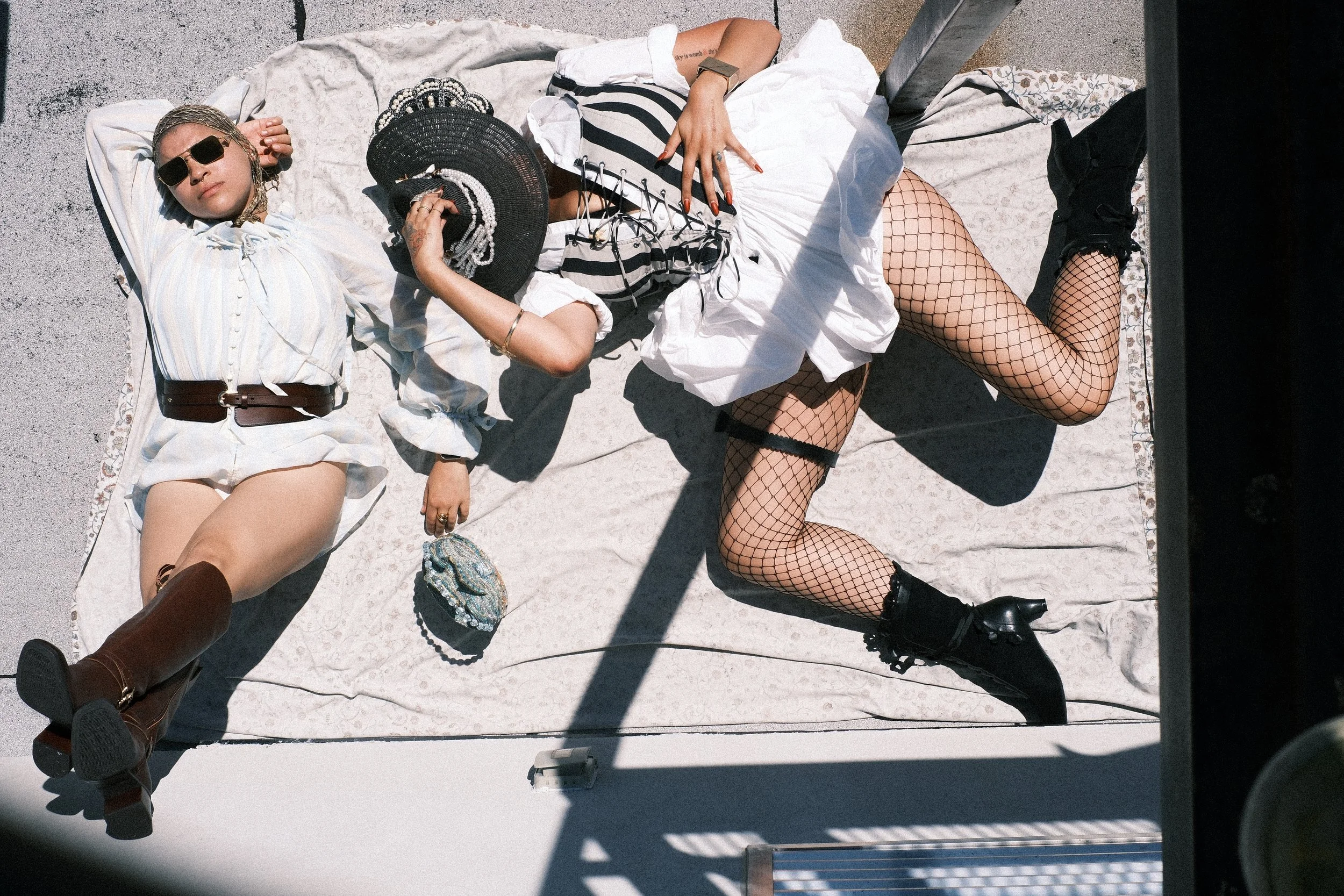 A woman lies on a white sheet on a concrete surface, wearing sunglasses, a white blouse, beige shorts, fishnet stockings, and black combat boots. She has a chain necklace and is holding a black hat with pearl decorations. There is a small, colorful b