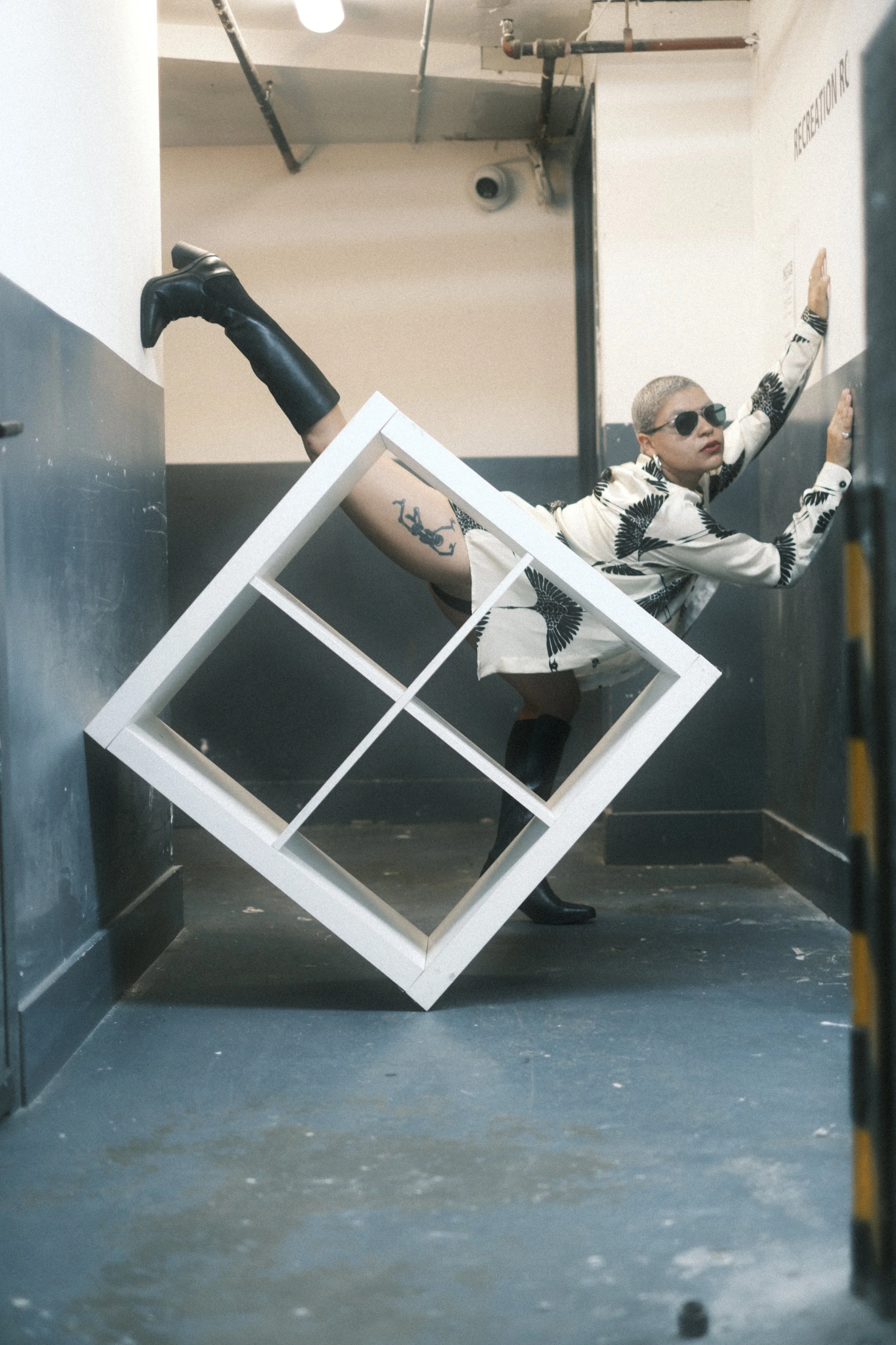 A woman with short blonde hair, wearing sunglasses, a patterned jacket, black thigh-high boots, and fishnet stockings, is posing against a wall in an industrial-style hallway, with her hand on the wall and one leg raised against the wall. There is a 
