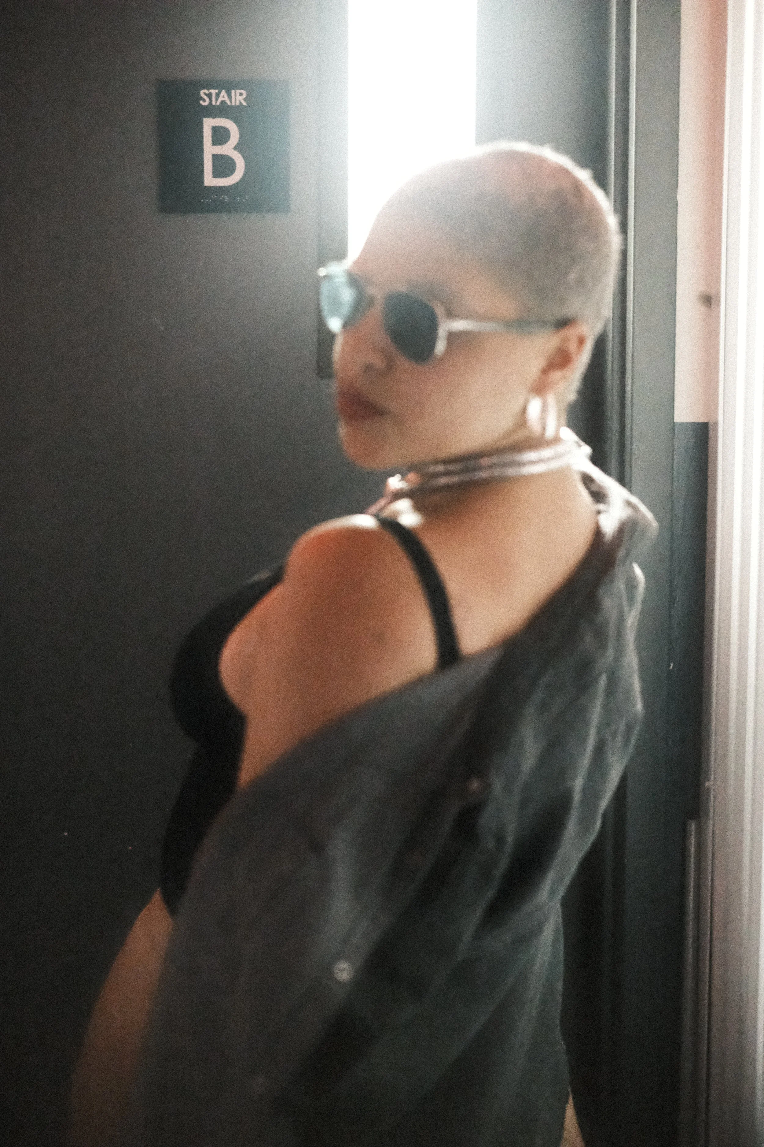A woman with short blonde hair wearing sunglasses, a sleeveless top with a black strap, and hoop earrings, standing in front of an elevator door with a sign indicating elevator B.