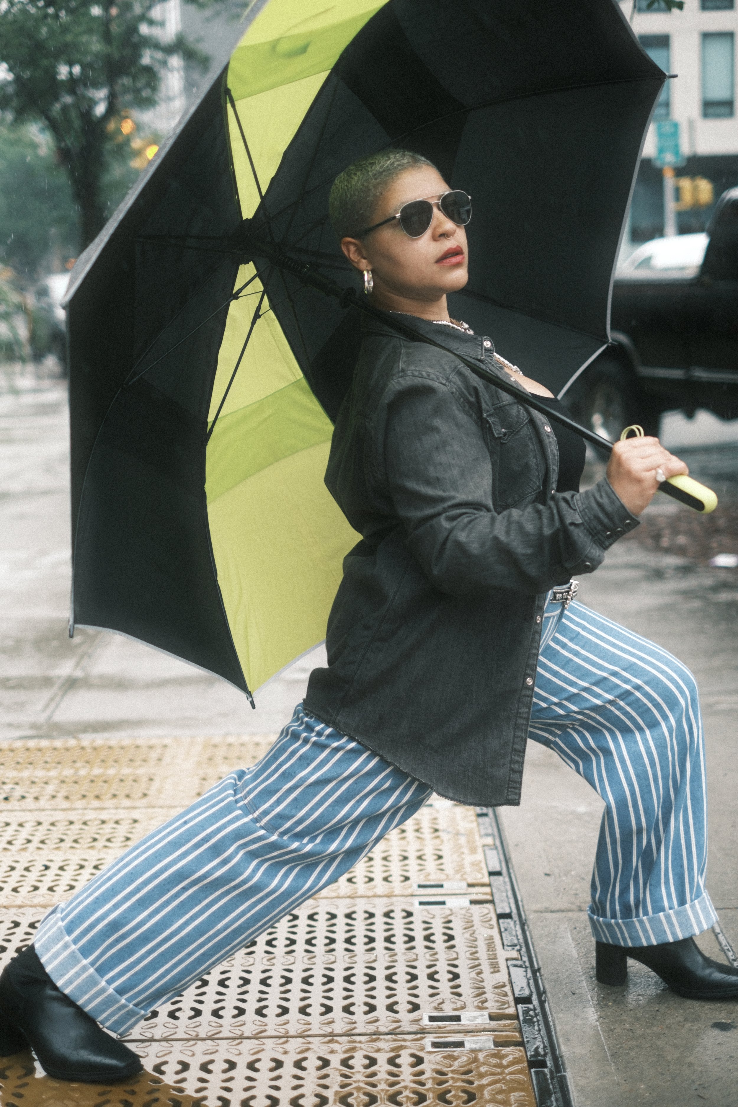 Woman with short hair, sunglasses, black leather jacket, and striped blue and white pants poses on a rainy city street holding a black and yellow umbrella.
