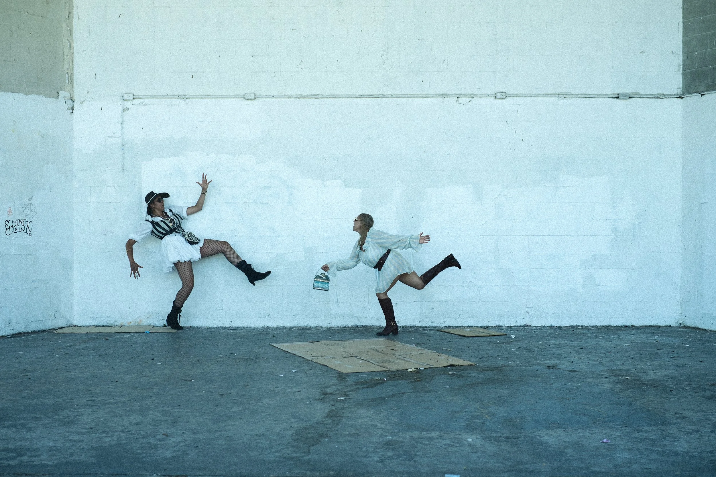 Two women dressed in eclectic outfits dance against a plain white wall in an empty, graffiti-stained space.