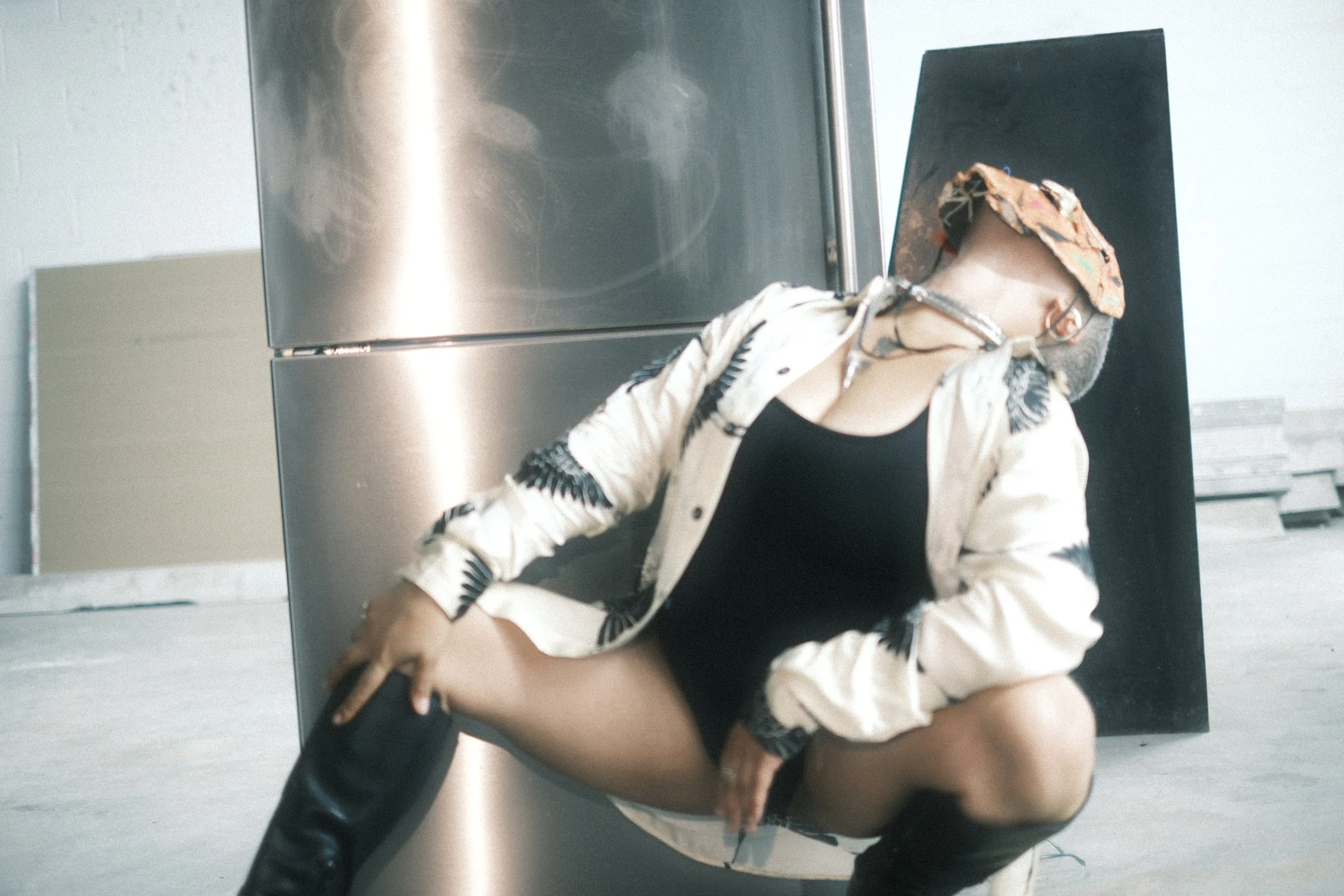 A person with a hat and glasses sitting in front of an elevator mirror, taking a selfie. The person is wearing a light-colored jacket with black and white patterns and black stockings.