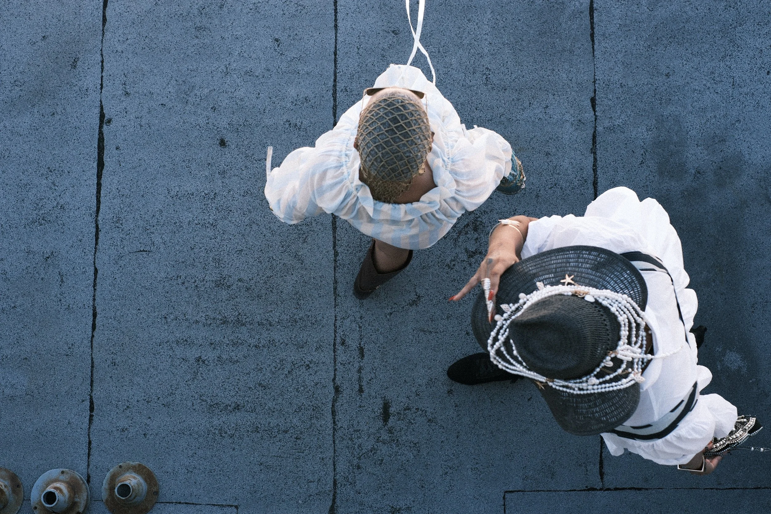 Overhead view of a person dressed in a white outfit and a wide-brimmed hat, standing on a paved surface along with a woman, with footwear and hats seen on the ground.