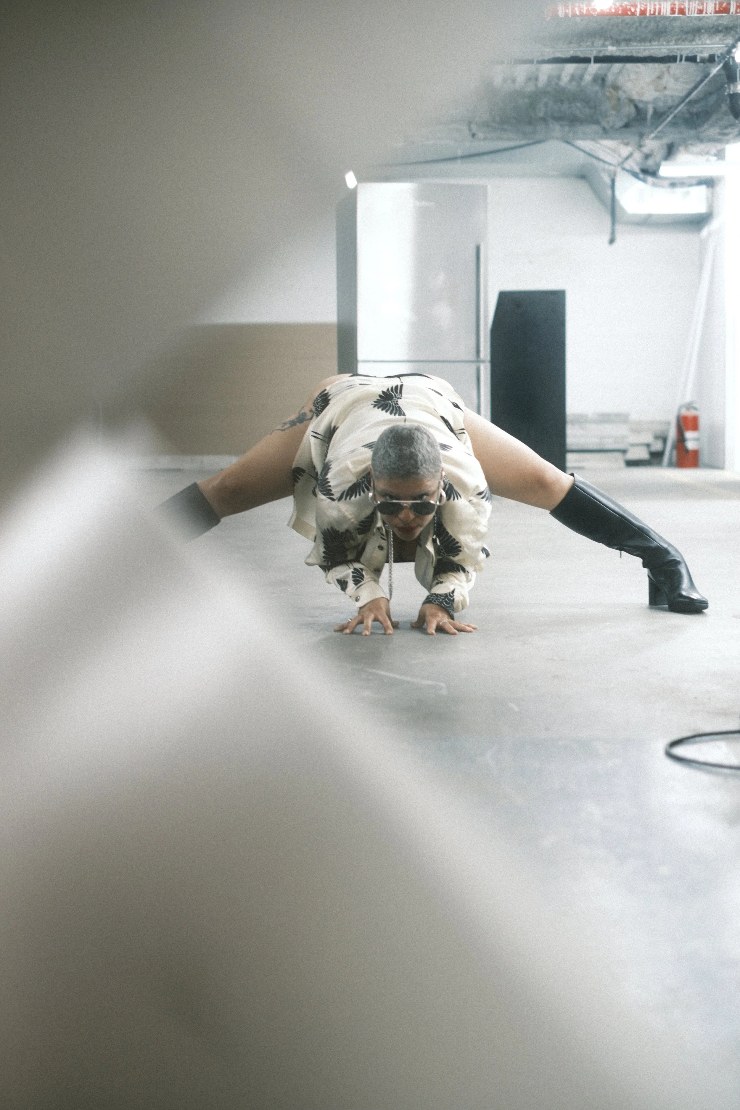 A person with short gray hair, wearing sunglasses and a patterned shirt, performing a yoga pose on the floor in an industrial-style space with concrete walls and a large refrigerator in the background.