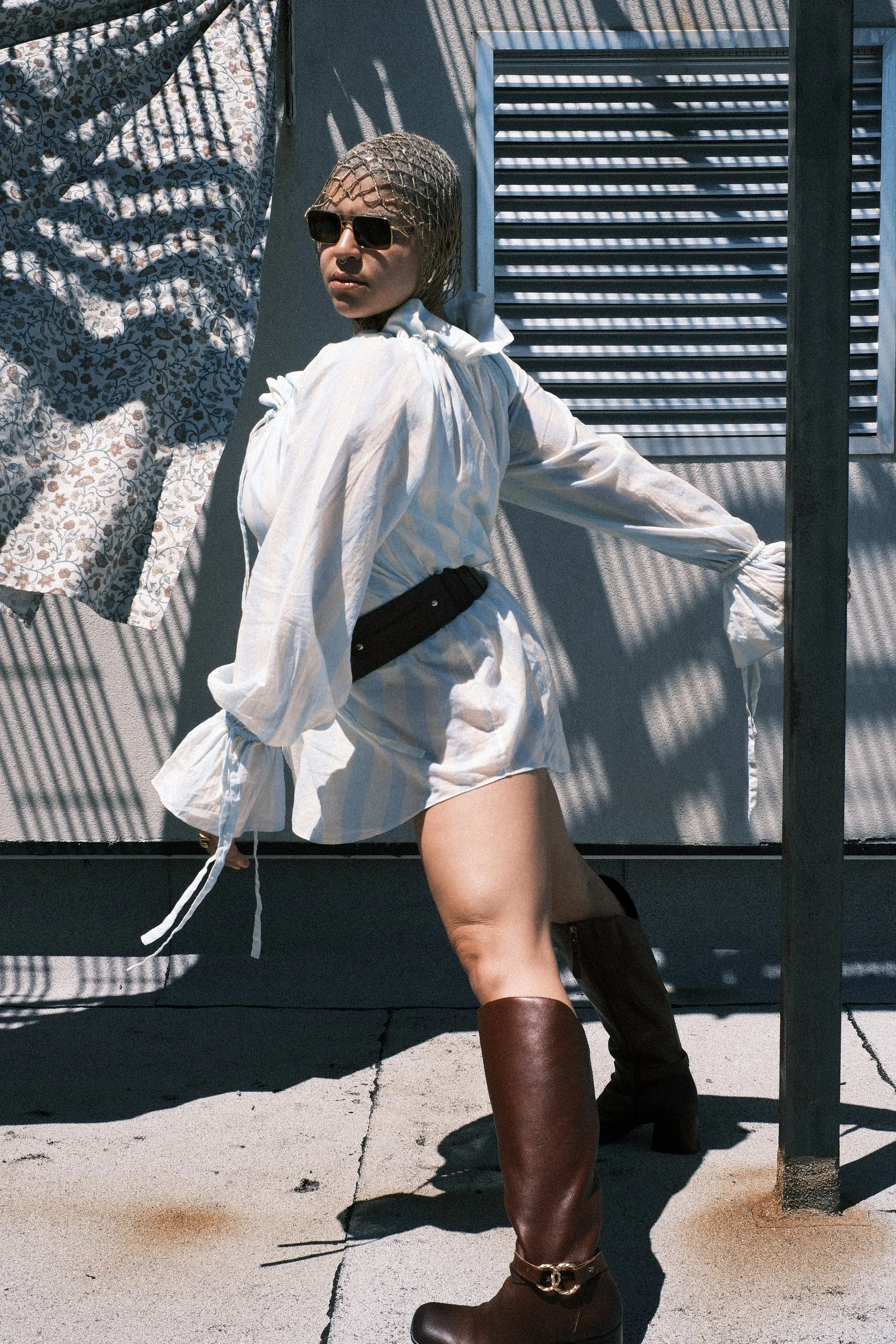 A woman wearing sunglasses, a netted head covering, and a white puffed-sleeve shirt poses outdoors against a textured wall with a floral fabric hanging to her left. She is wearing brown knee-high boots and a black belt.