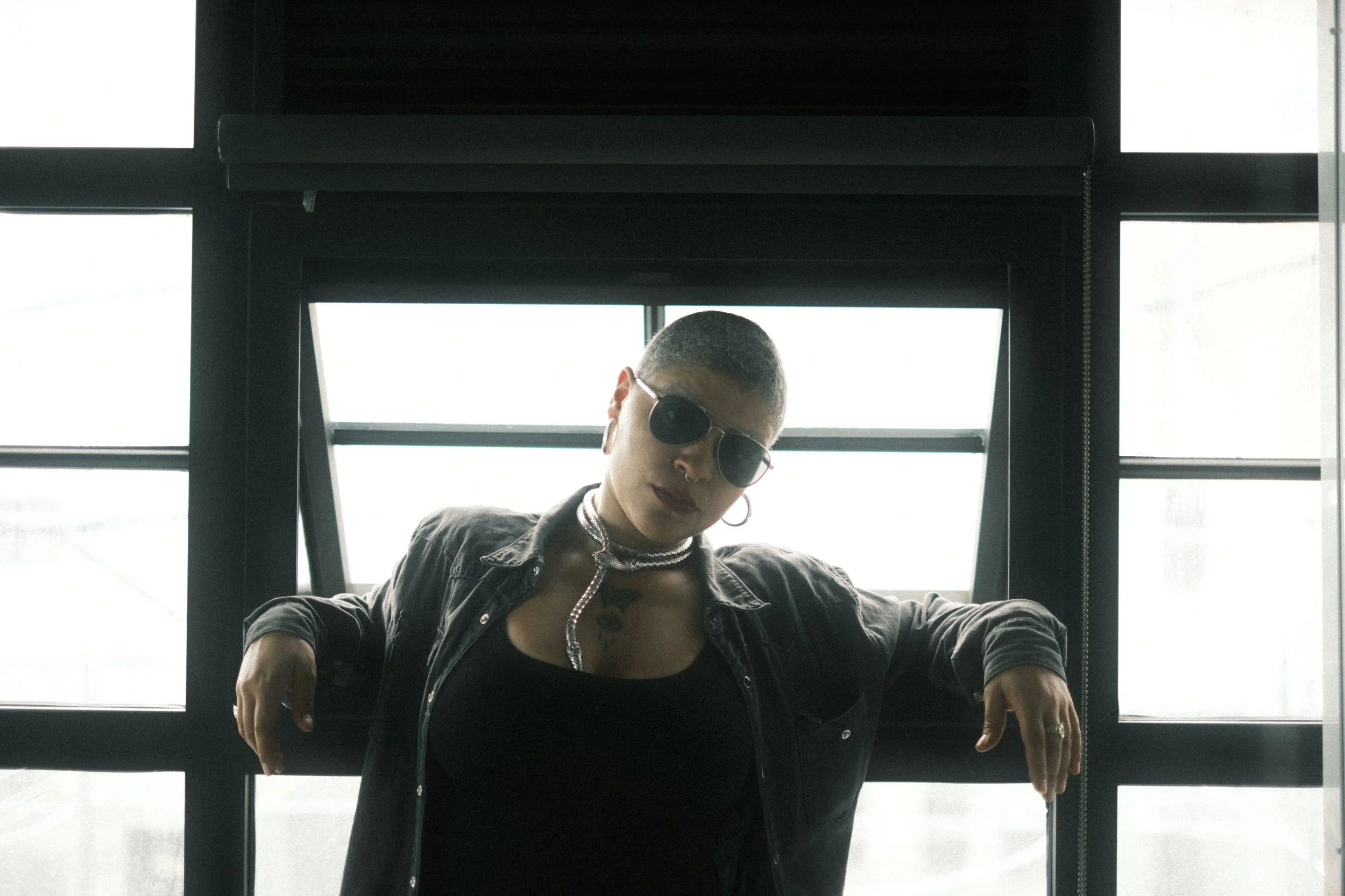 A person with short hair and sunglasses, wearing a black jacket and necklace, leaning over a railing in front of large windows in a modern building.