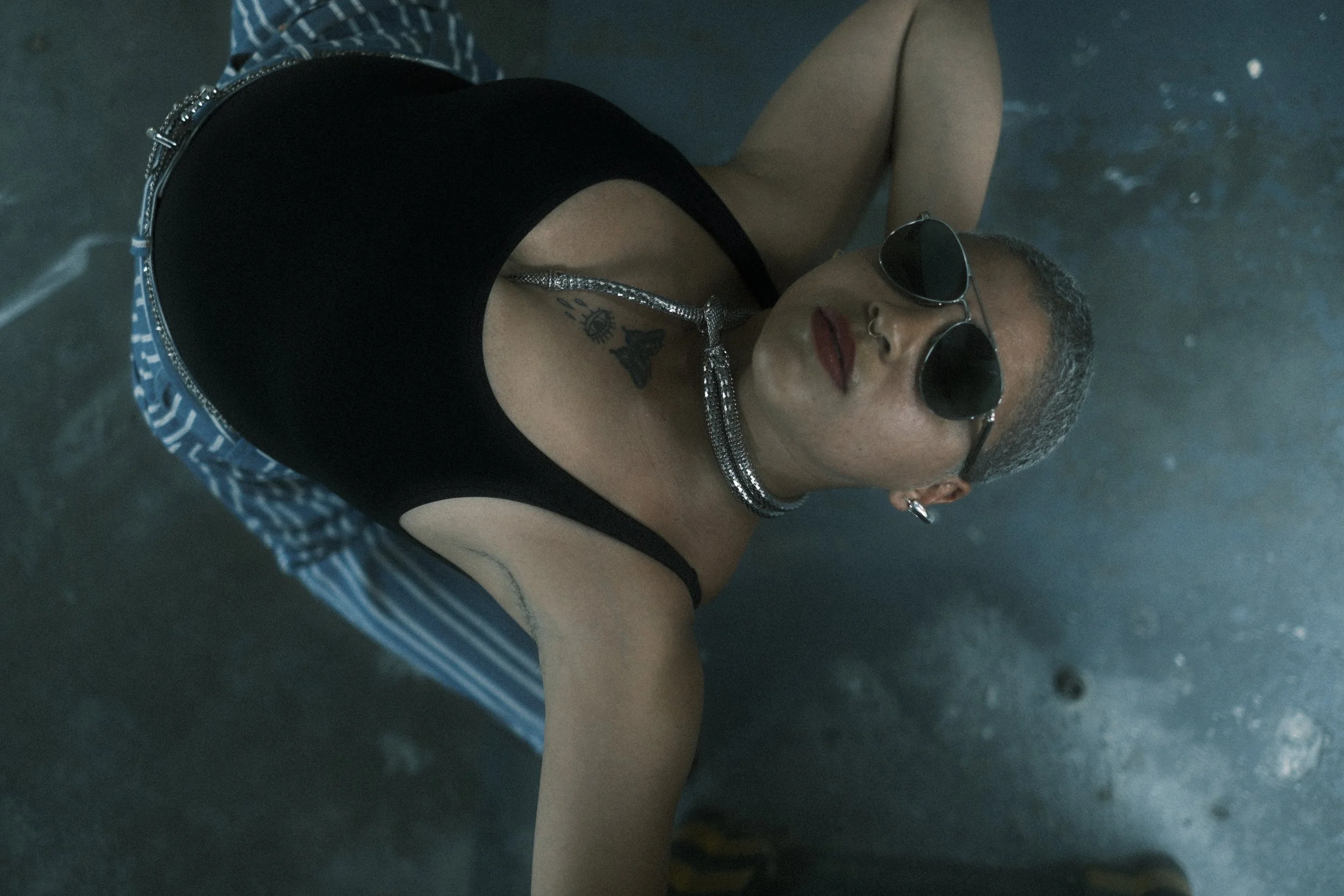 A woman with short gray hair and dark sunglasses lying on the ground, wearing a black sleeveless top, a long silver necklace, and sporting tattoos on her shoulder and chest.