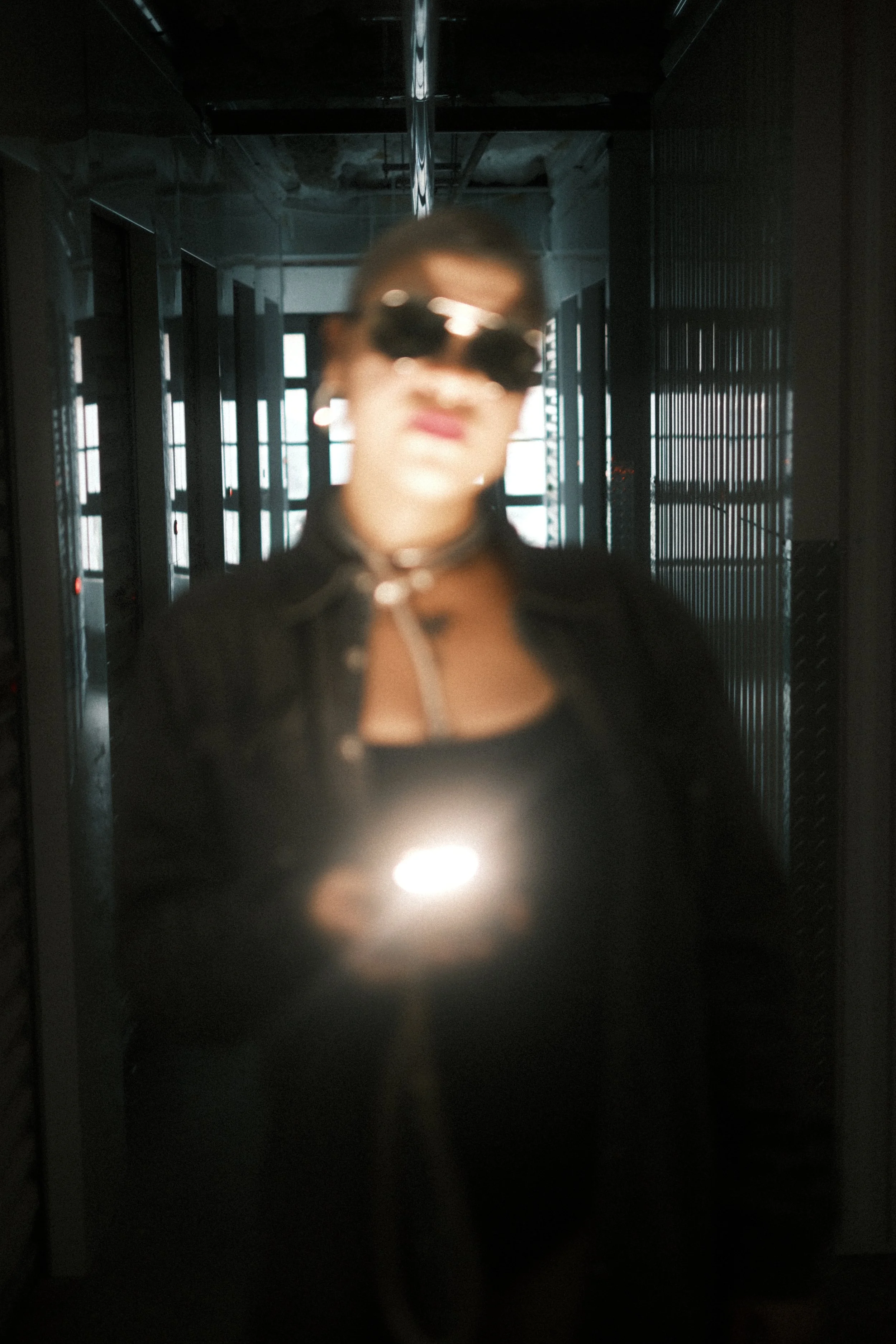 A blurry person wearing sunglasses and a choker necklace, standing in front of a dark background with mirrored surfaces and some light coming through windows.
