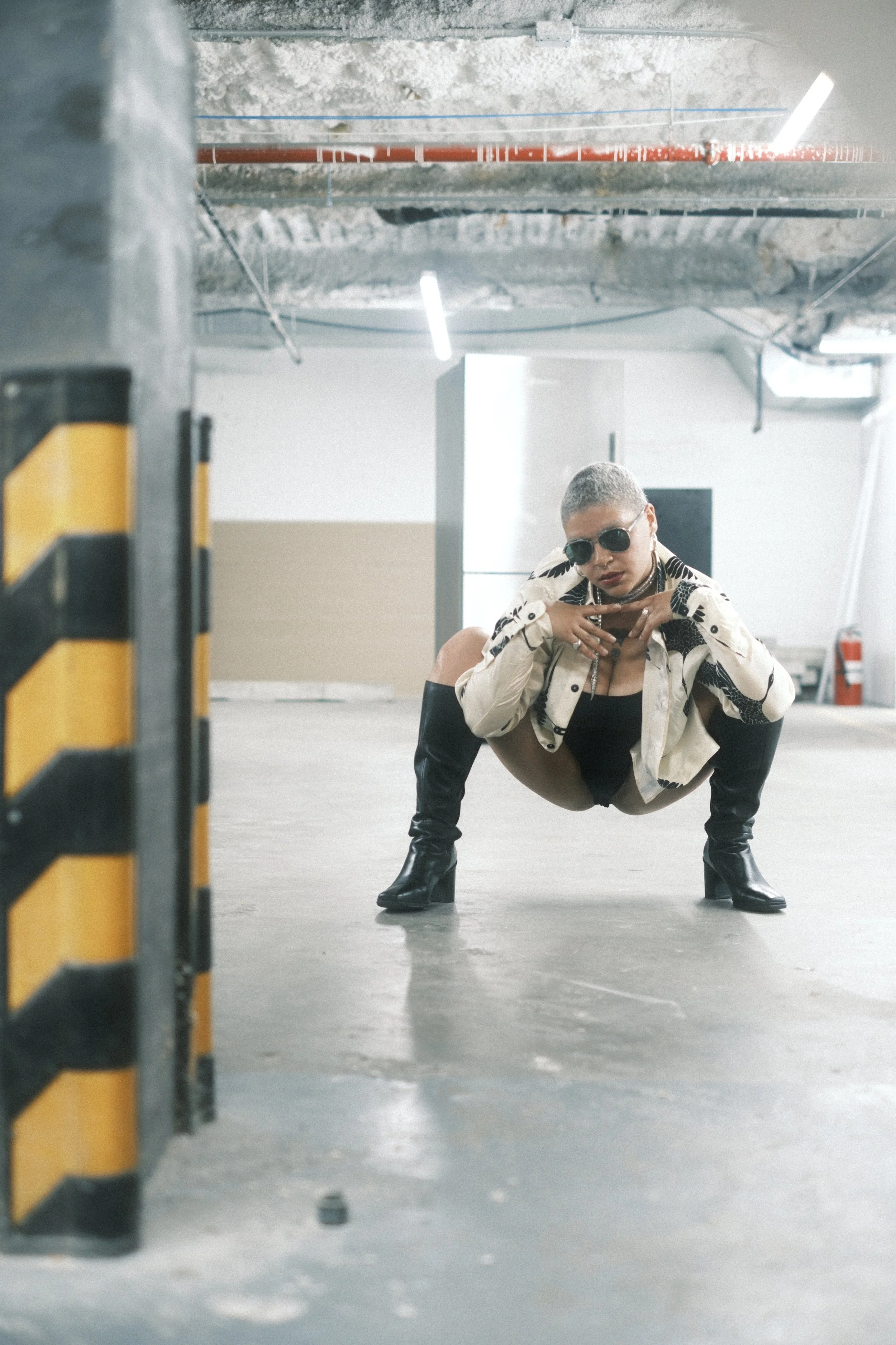 A person with short hair, wearing black sunglasses, a patterned beige jacket, black boots, and shorts, is crouching in an industrial parking garage.