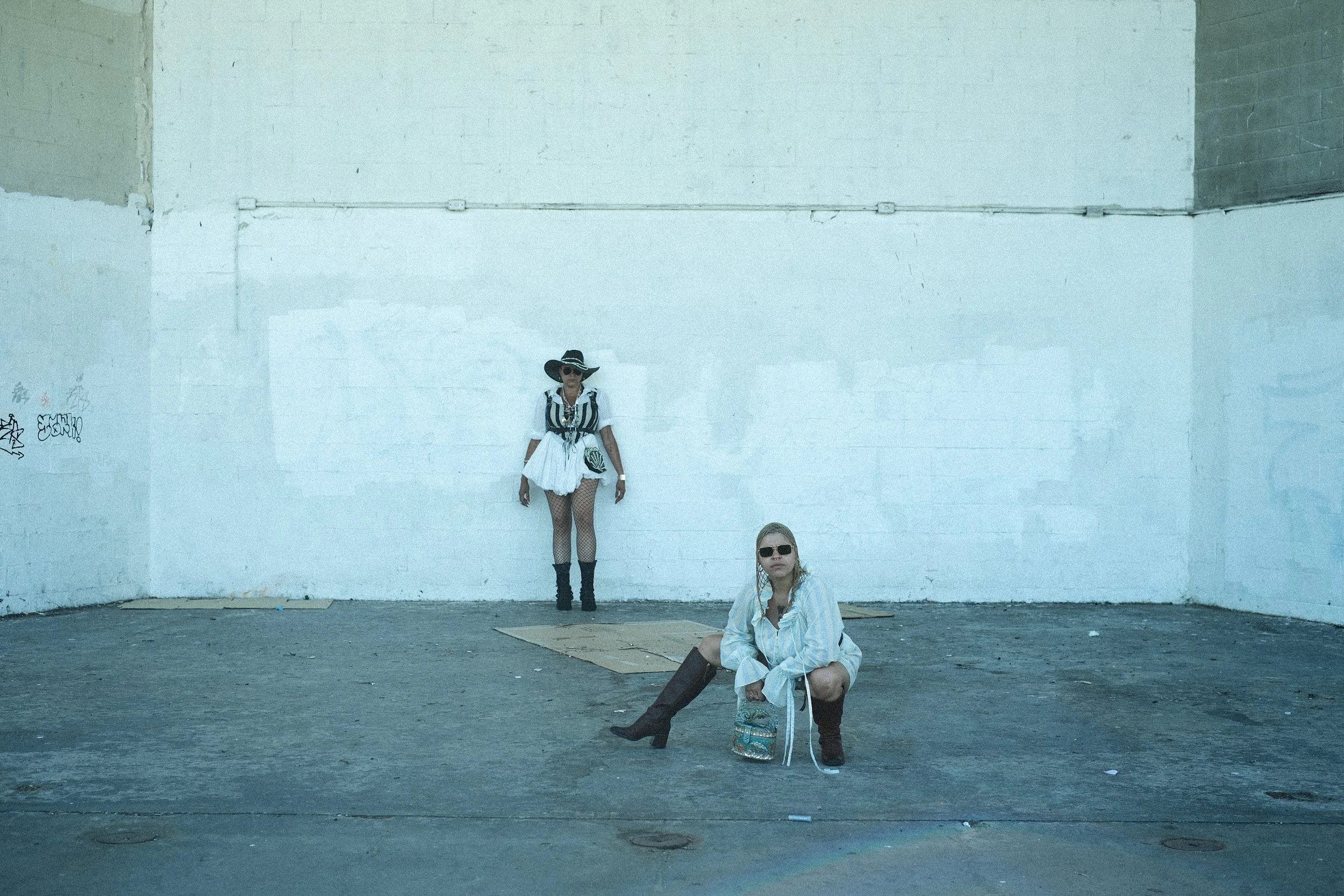 Two women with sunglasses and fashionable clothing in an industrial outdoor space with a large white wall. One woman is sitting on the ground with her legs crossed, and another is standing against the wall wearing a hat and fishnet stockings.